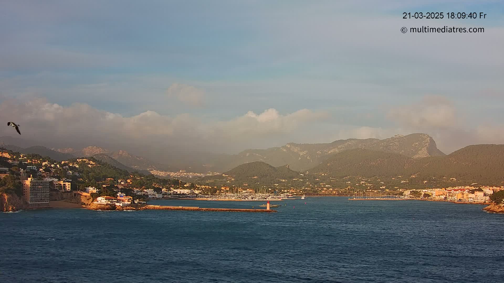 A coastal town nestled against mountains under a partly cloudy sky, with a calm sea in the foreground and a bird in flight.