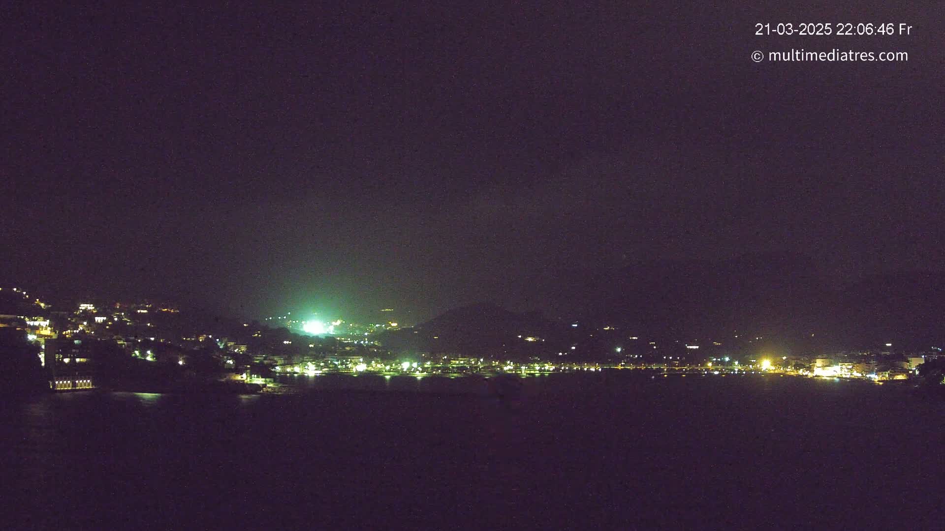 A nighttime, dark view of a coastal town with its lights illuminating the hillsides and waterfront under a mostly clear sky.
