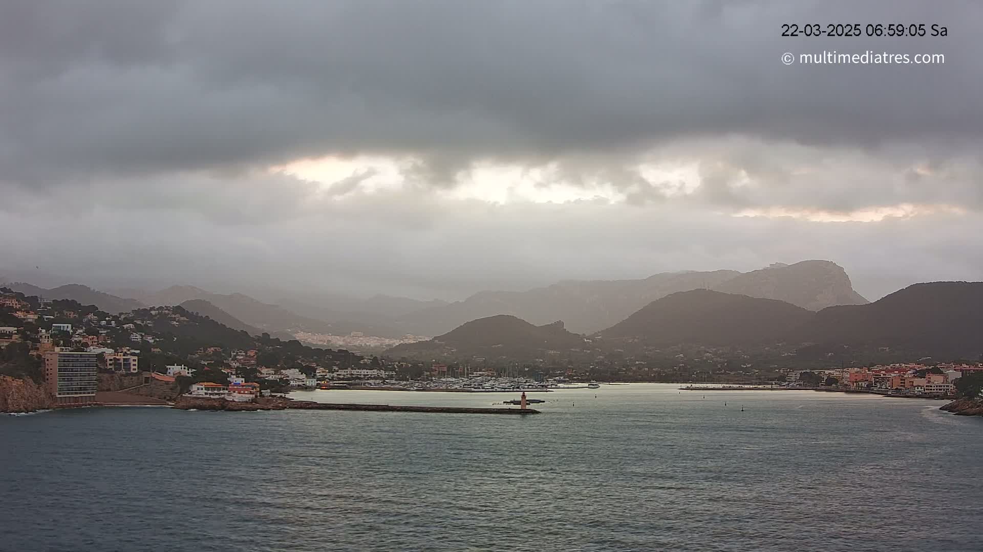 A coastal town nestled between mountains under a cloudy, overcast sky sits on a calm body of water with a harbor full of boats.