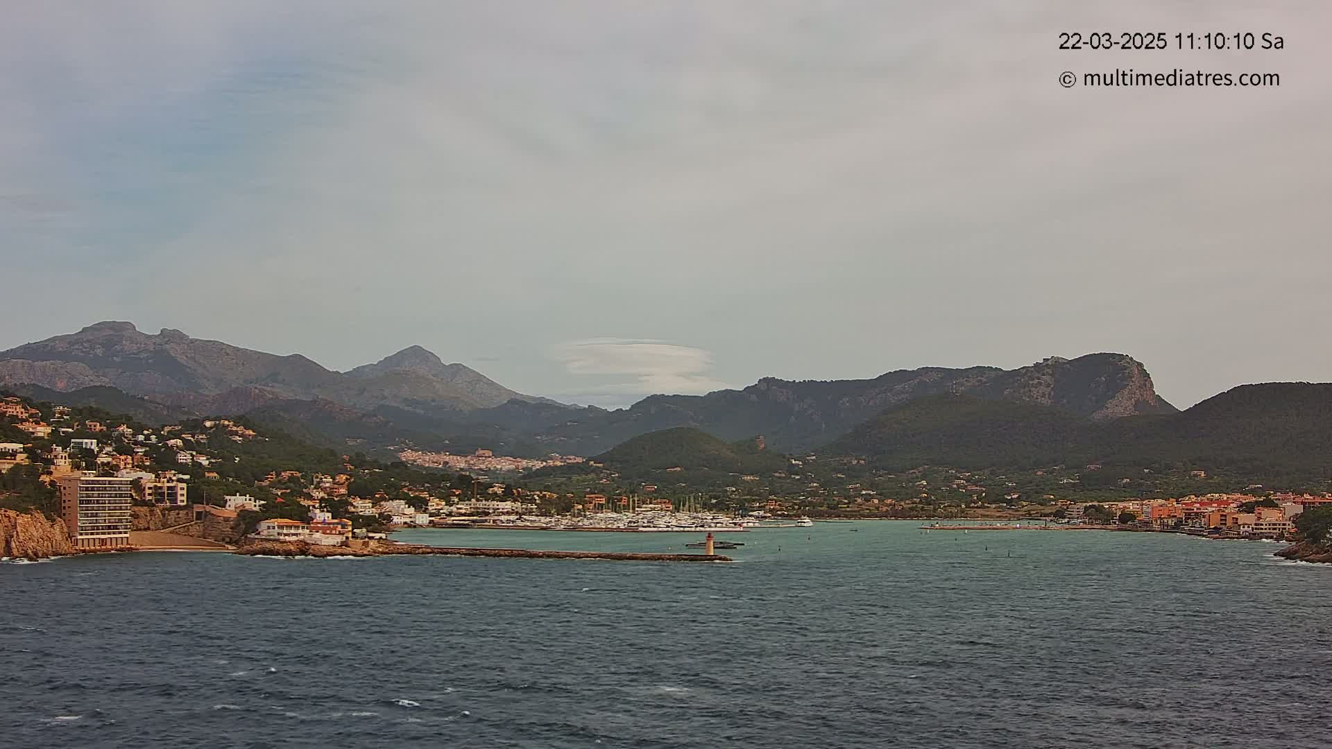 A coastal town nestled between mountains, with a marina filled with boats, under a mostly cloudy sky.