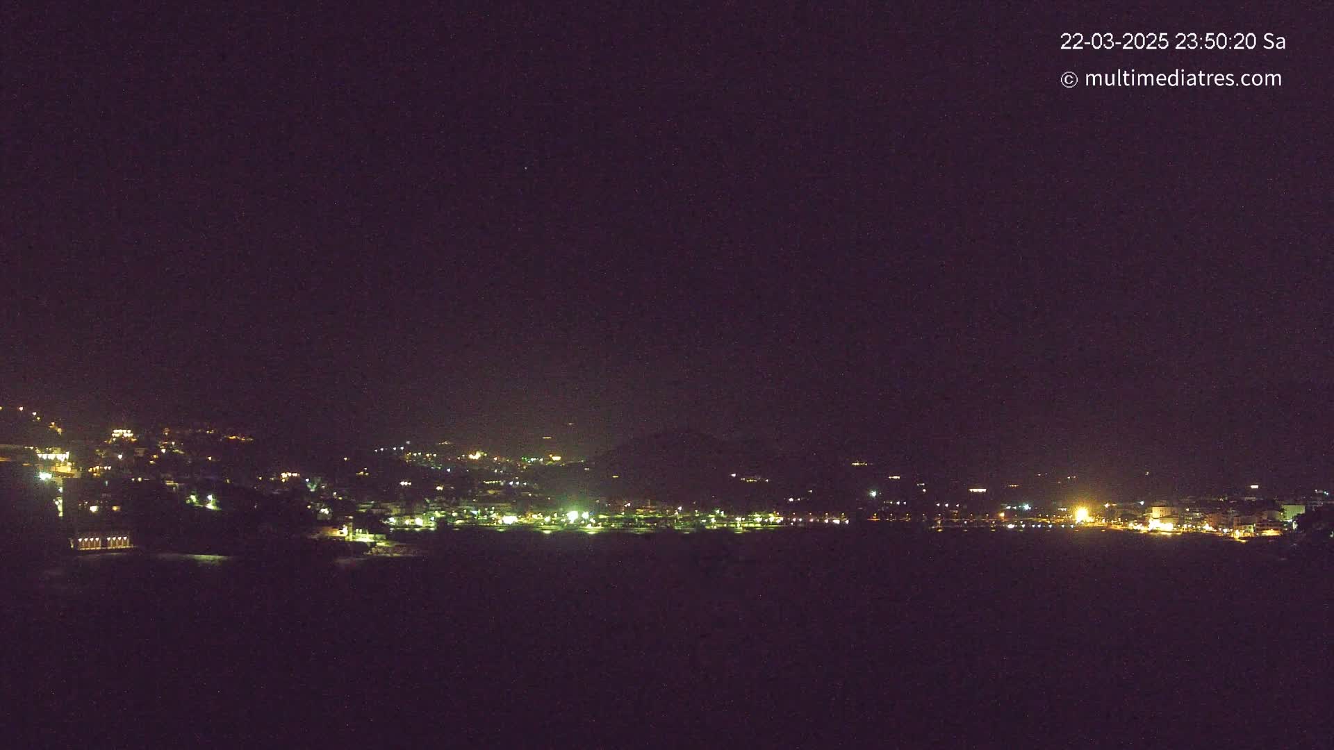 A nighttime long shot shows a coastal town with scattered lights on a hill and along a dark body of water under a clear, dark sky.