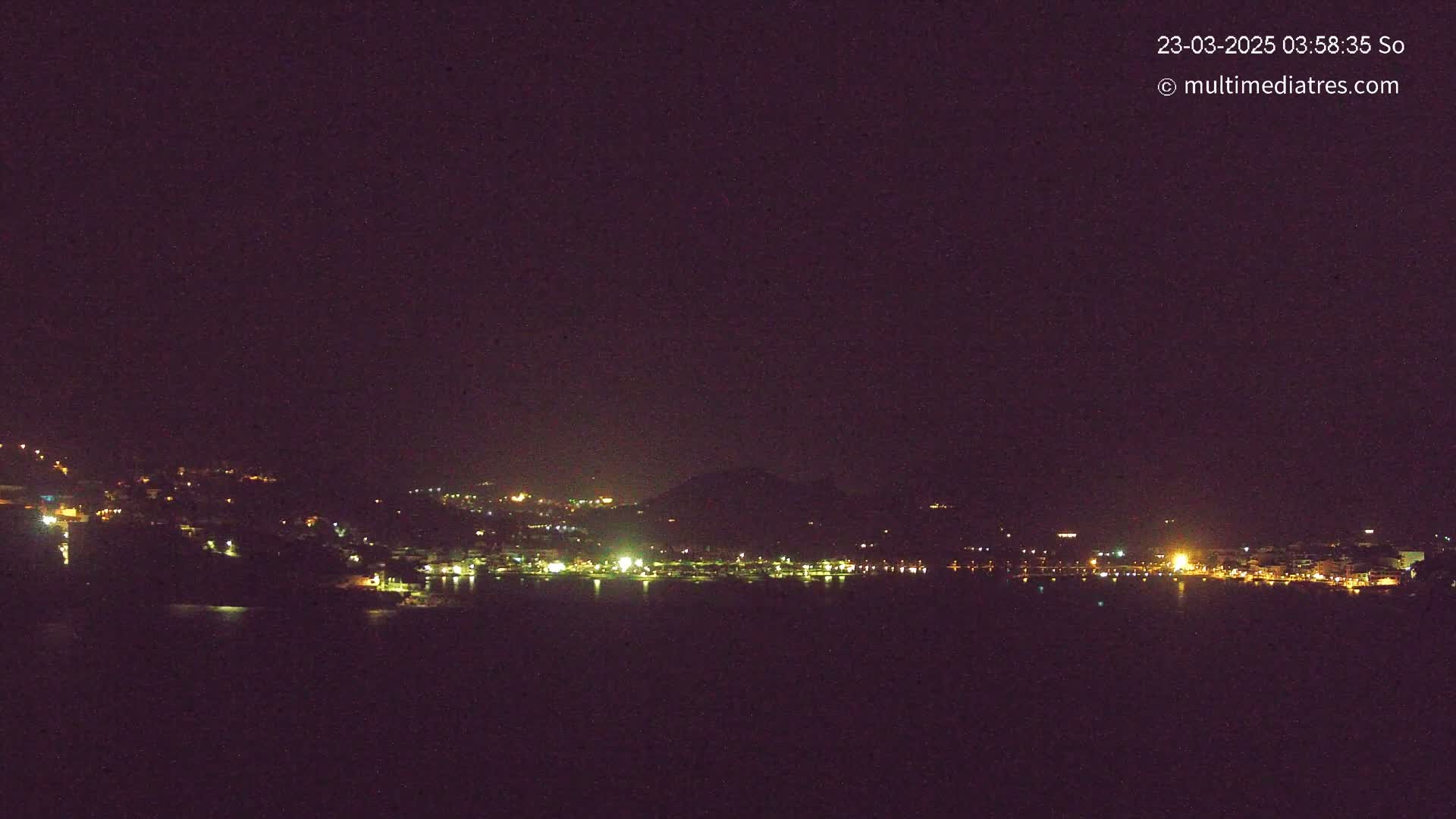 A nighttime view shows a dark, calm body of water reflecting the faint lights of a town nestled against a dark hillside under a clear, starless sky.