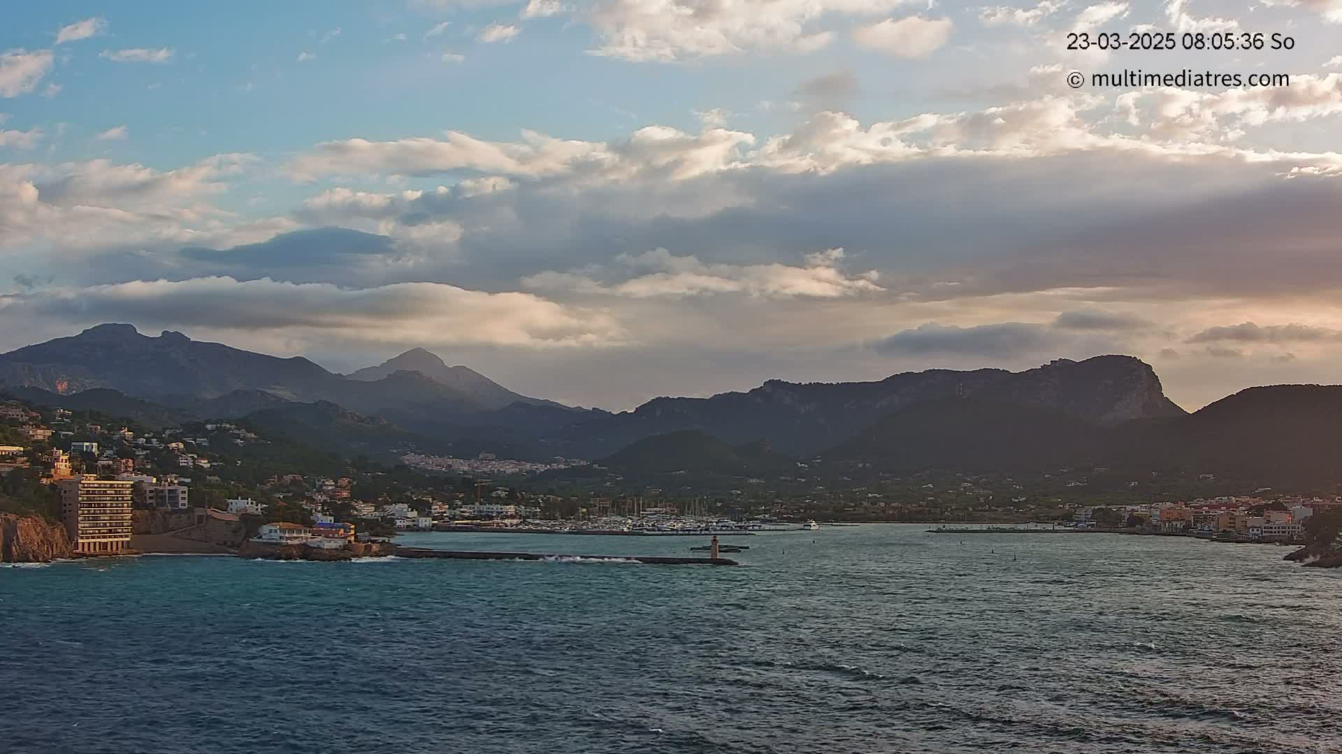 A partly cloudy sky overlooks a coastal town nestled between mountains and a calm sea with a harbor filled with boats.