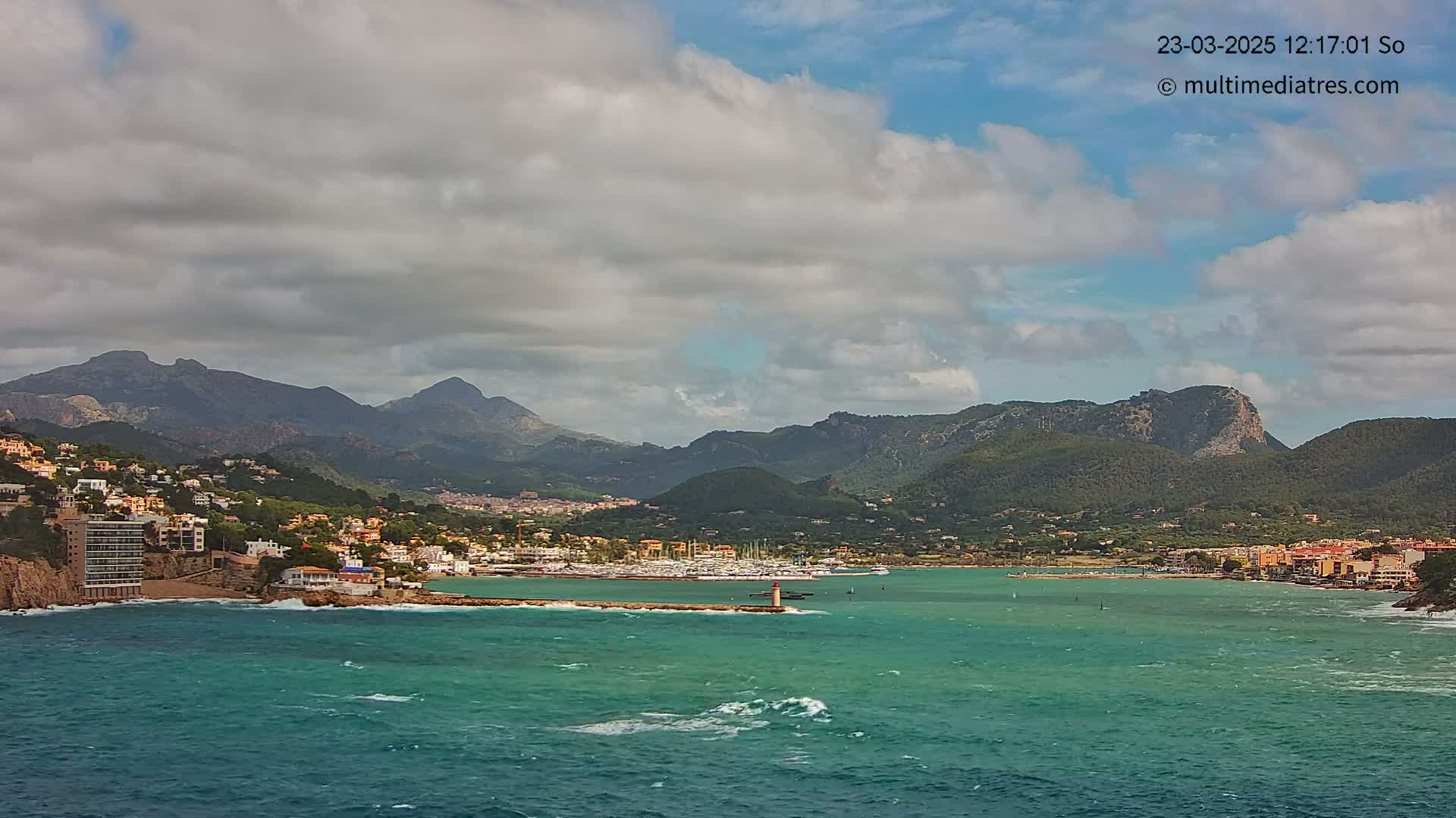A partly cloudy day reveals a coastal town nestled between a turquoise sea and a mountain range, with a small lighthouse marking a harbor filled with boats.