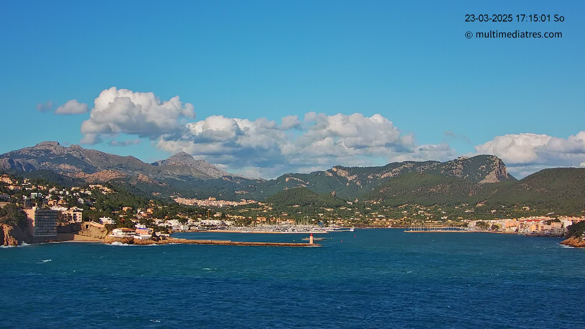 A coastal town nestled against a mountain range under a partly cloudy blue sky, with a calm sea in the foreground.