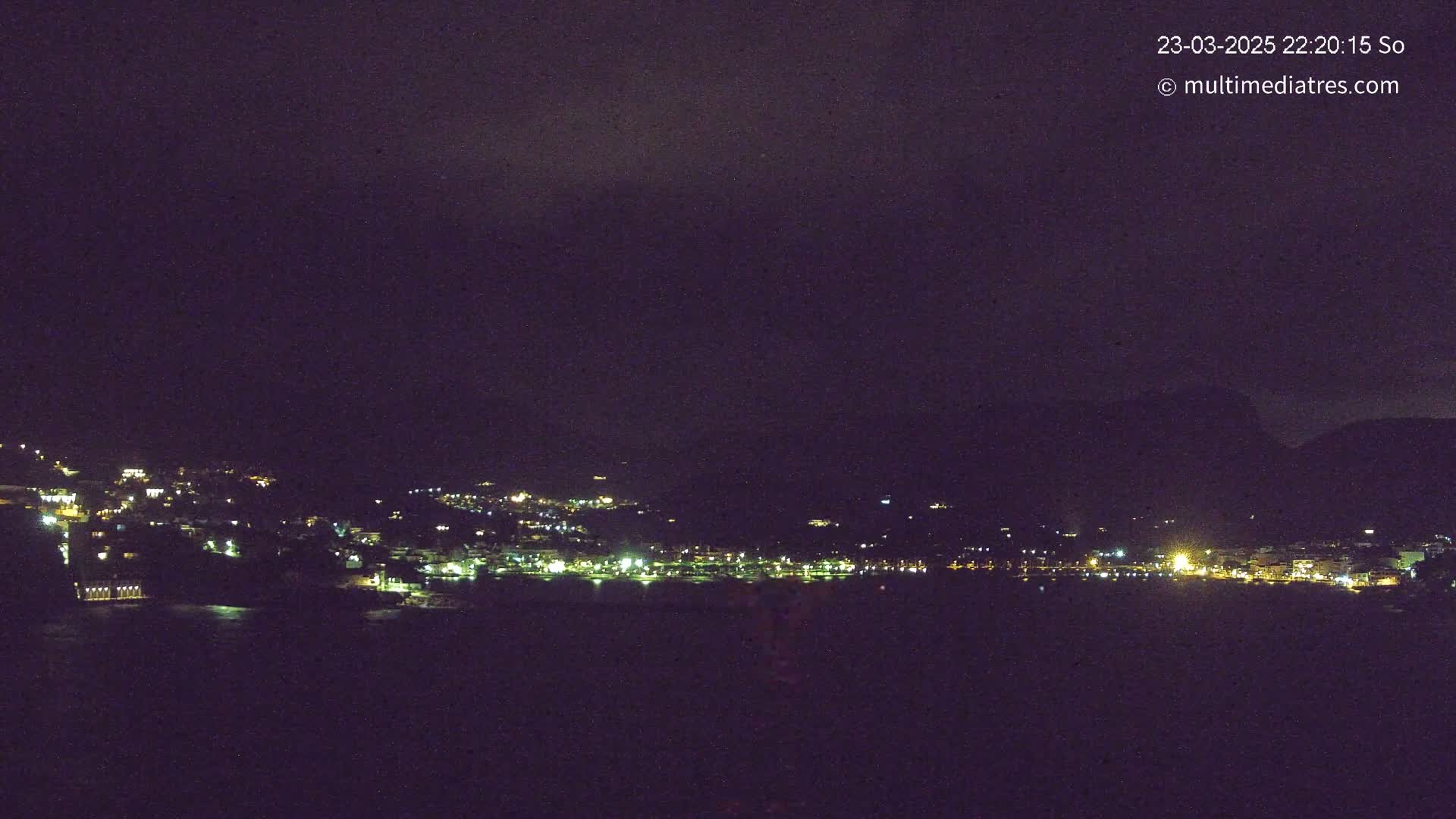 A nighttime, dark, coastal town with faintly visible lights is seen from across a dark body of water under a cloudy sky.