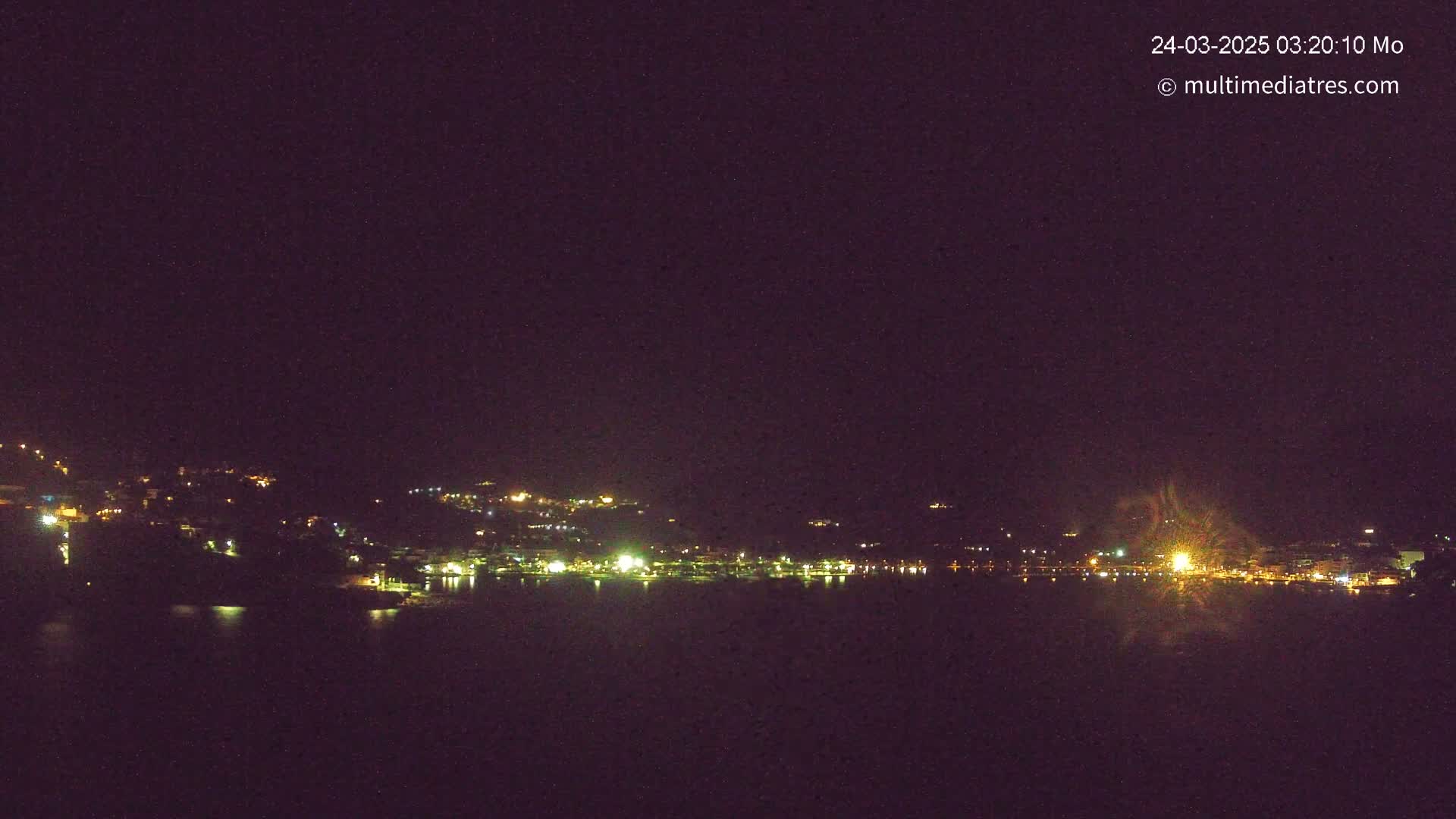 A nighttime long shot of a town's lights along a dark body of water, under a clear, dark sky.