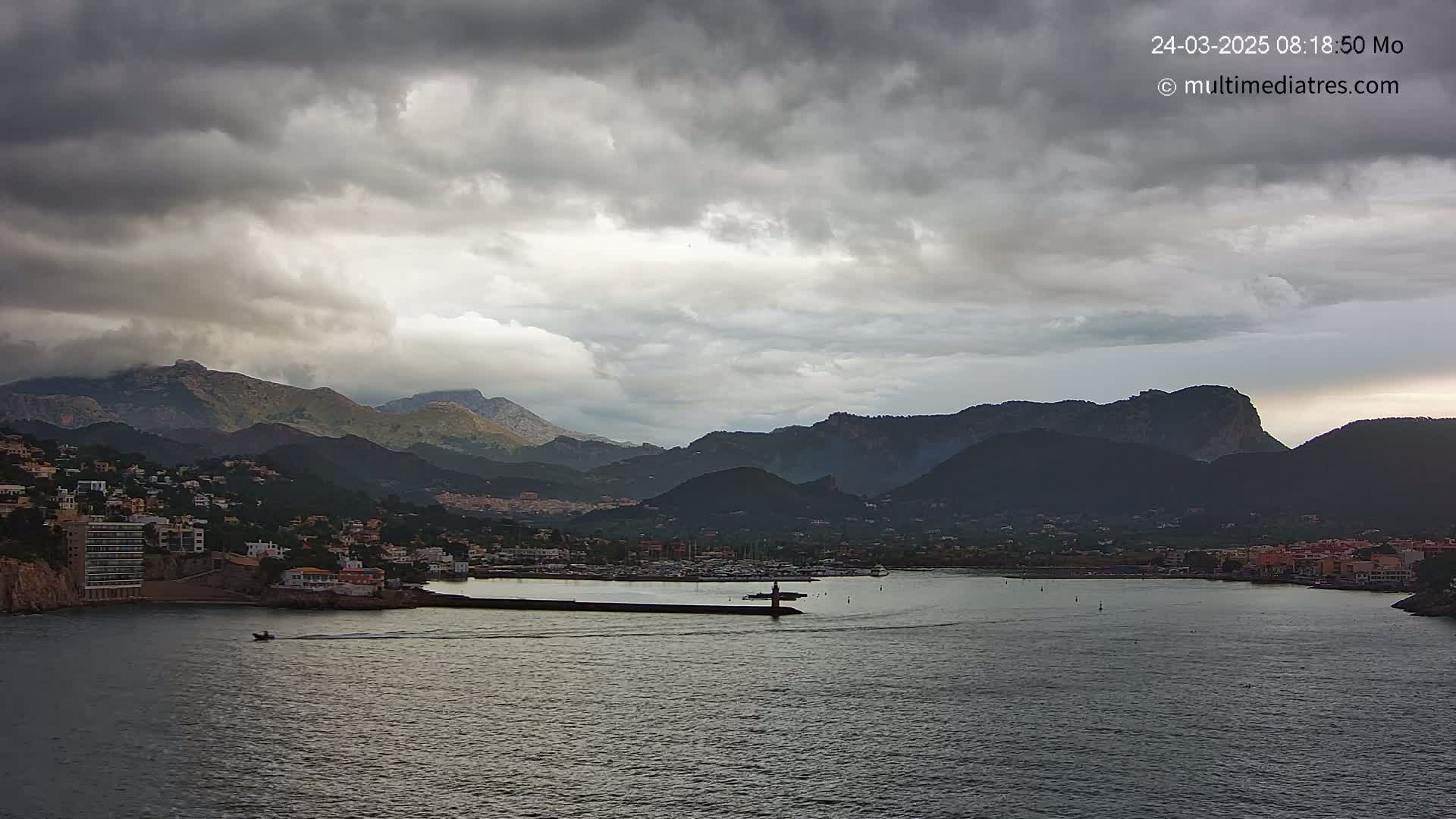 A coastal town nestled between dark mountains under a cloudy sky sits on a calm body of water.