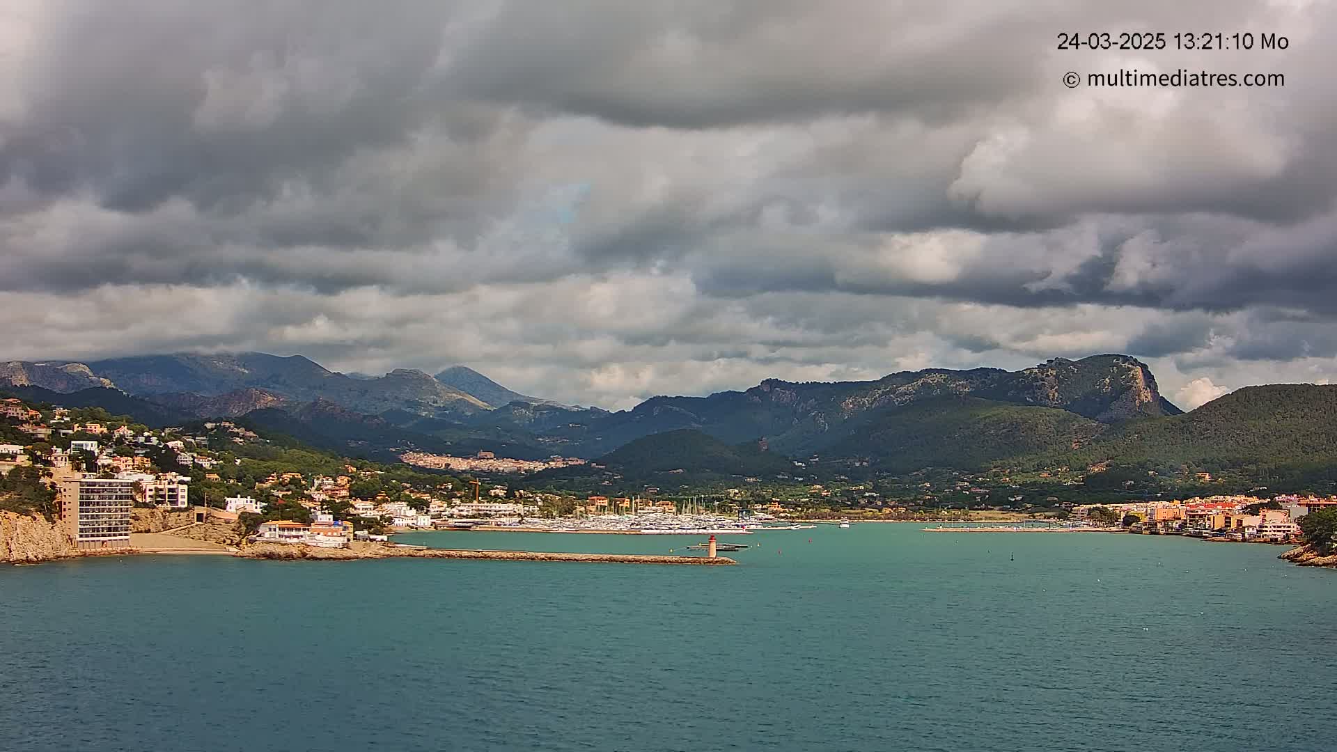 A coastal town nestled against a mountainous backdrop under a cloudy sky, with a harbor filled with boats visible in the mid-ground.