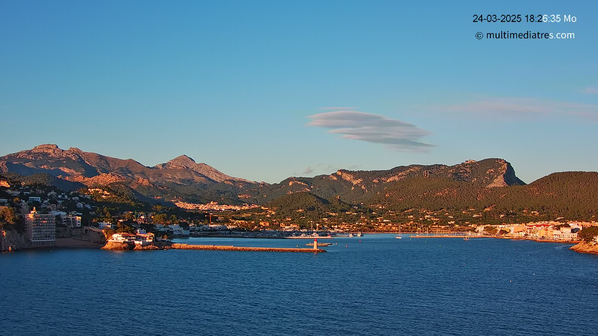 A coastal town nestled at the base of mountains under a clear blue sky with a small, wispy cloud.