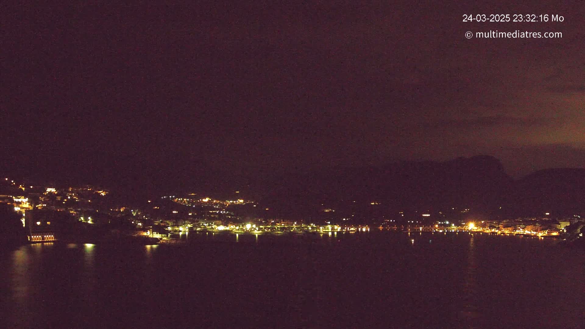 A nighttime view of a coastal town illuminated by lights, with dark hills in the background and calm water in the foreground under a dark, possibly cloudy sky.