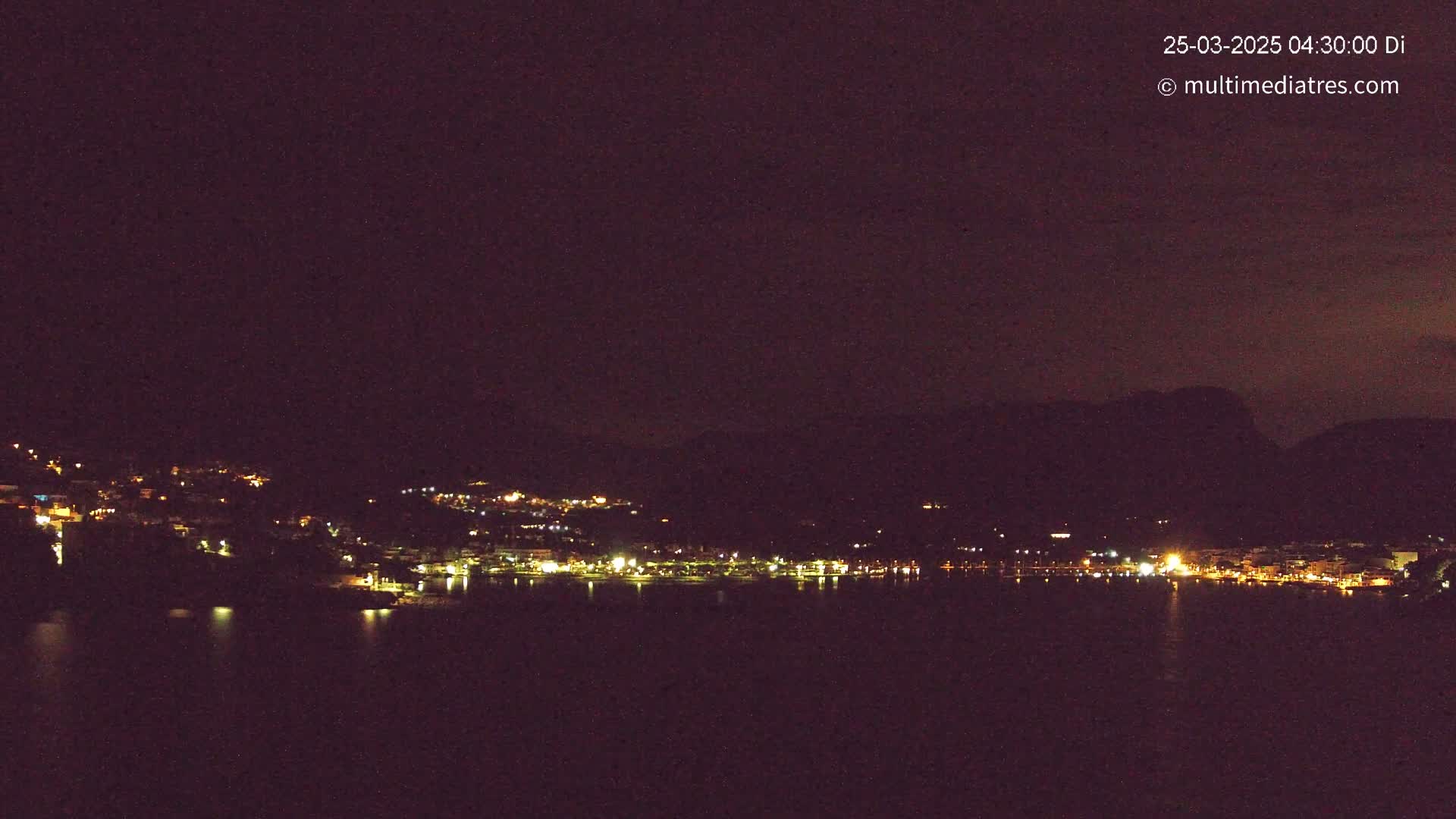 A nighttime view shows a dark, hilly coastline with scattered lights from buildings and a dark body of water in the foreground under a clear, dark sky.