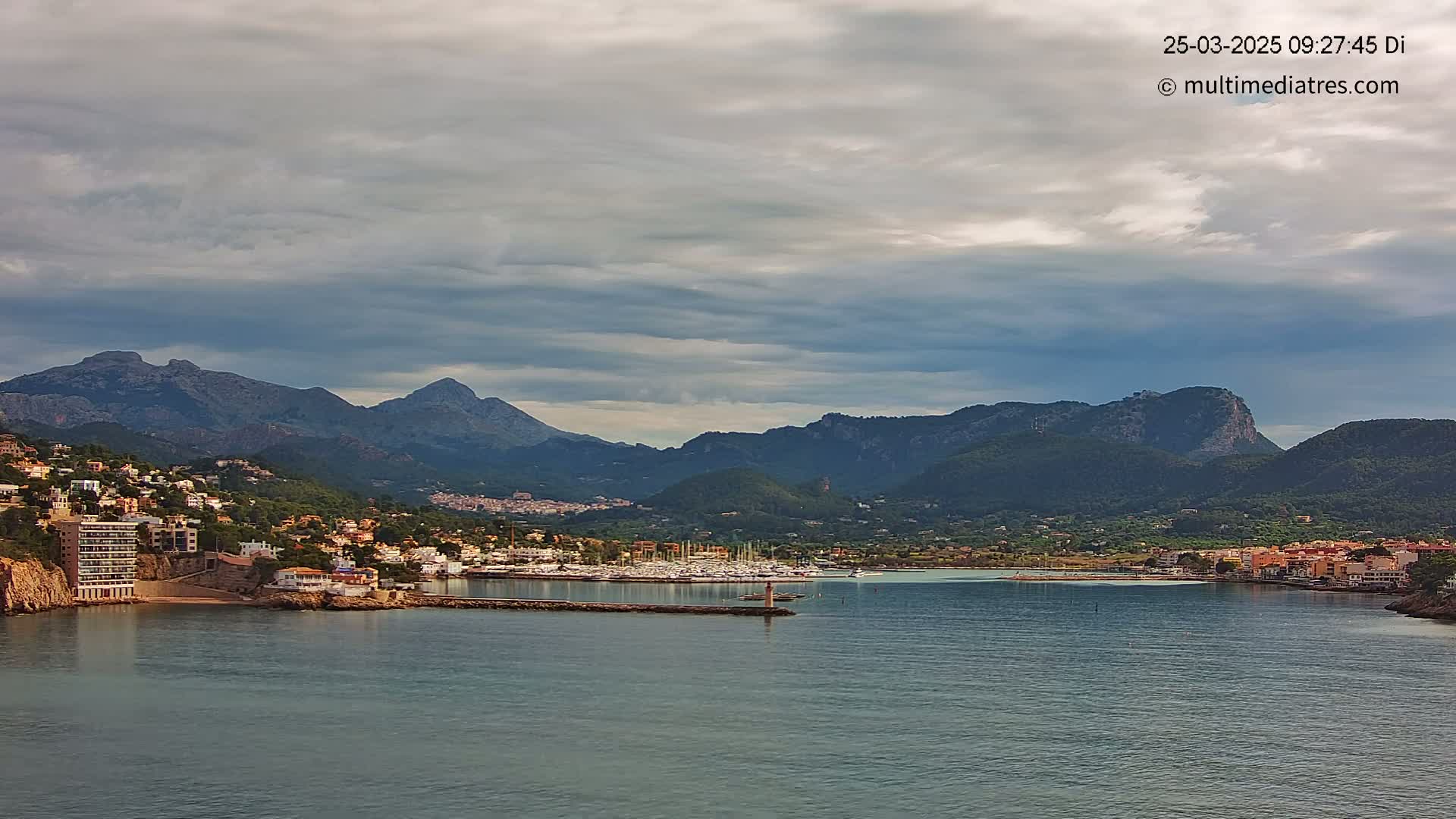 A coastal town nestled between mountains under a cloudy sky, with a marina filled with boats in the foreground.