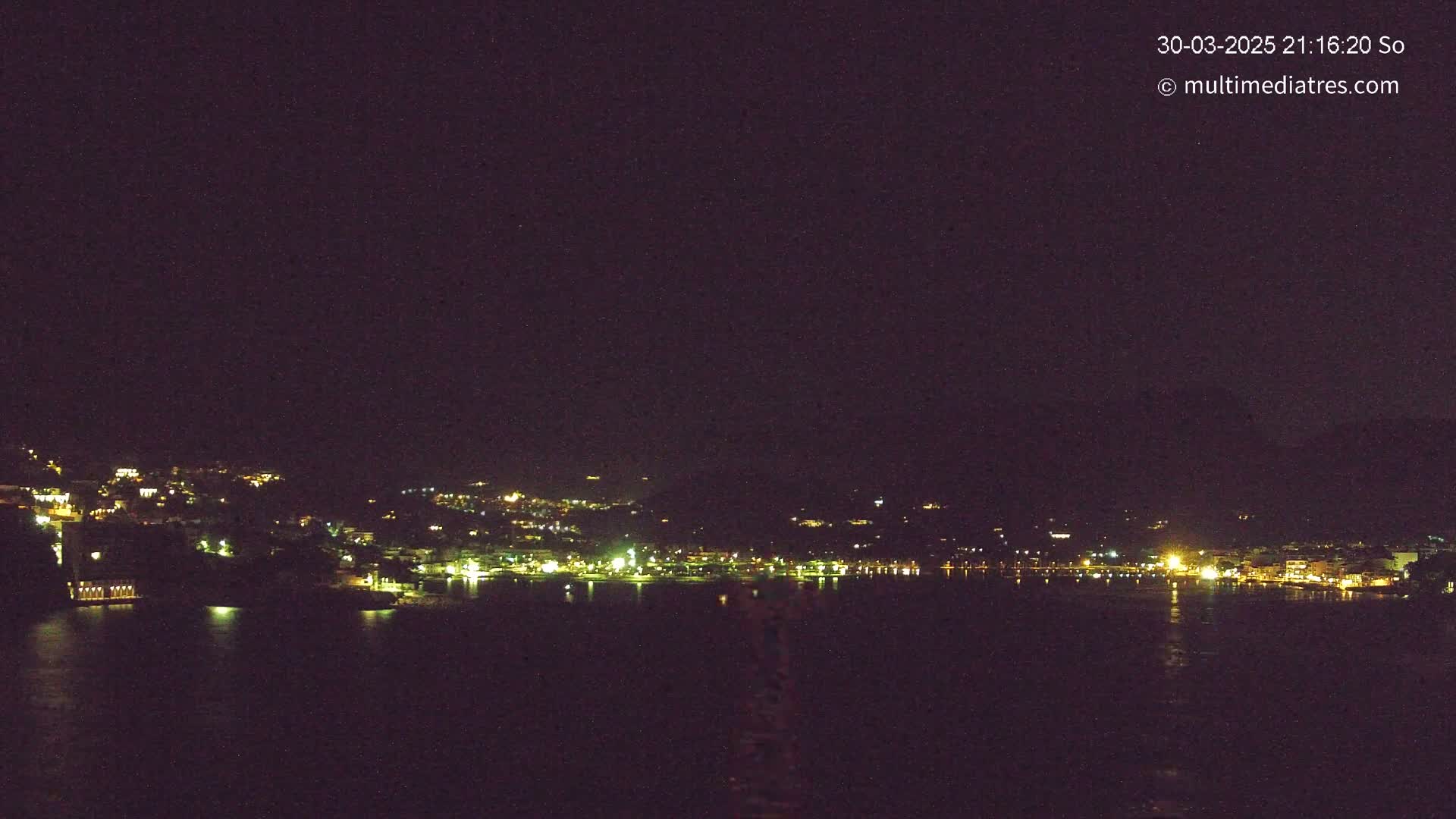 A nighttime view of a coastal town with its lights illuminating the waterfront and hillsides under a clear, dark sky.
