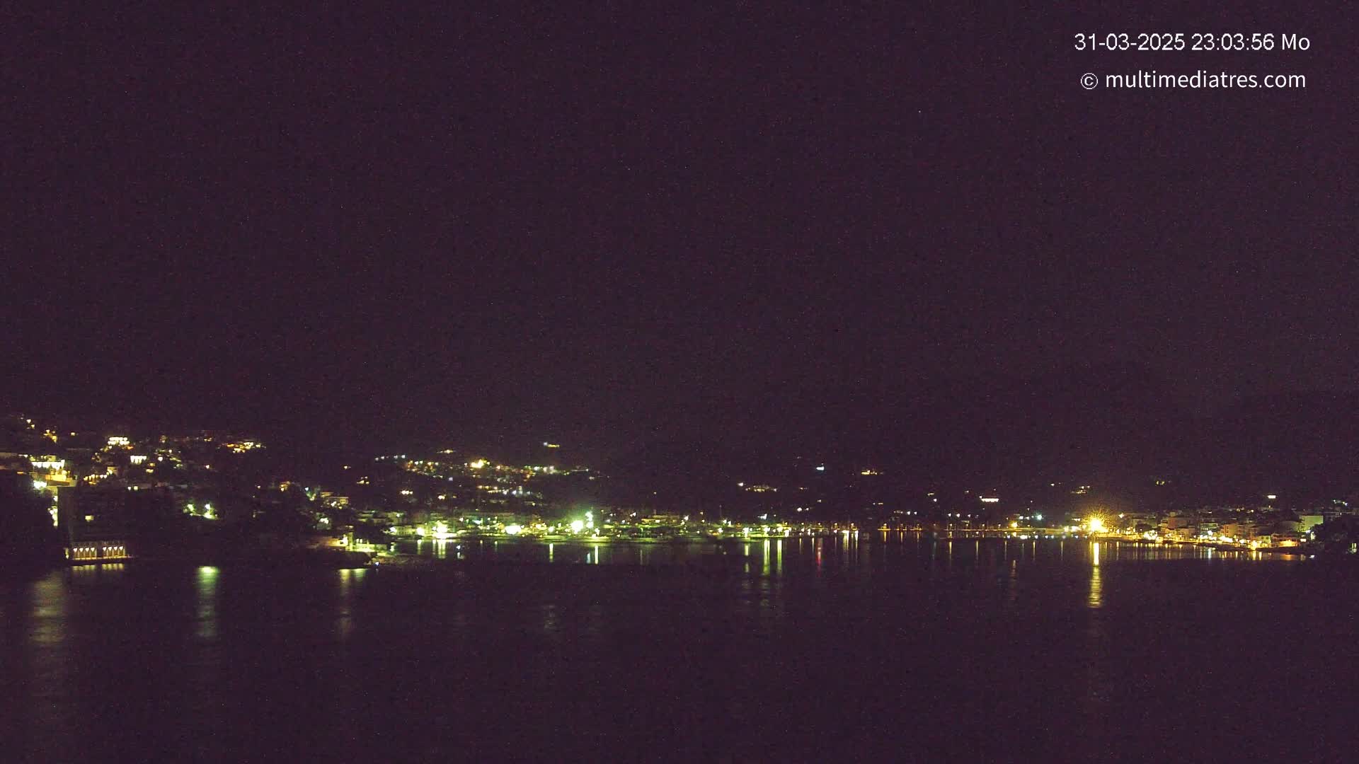 A nighttime view of a coastal town's lights reflected on a calm, dark body of water under a clear, starlit sky.