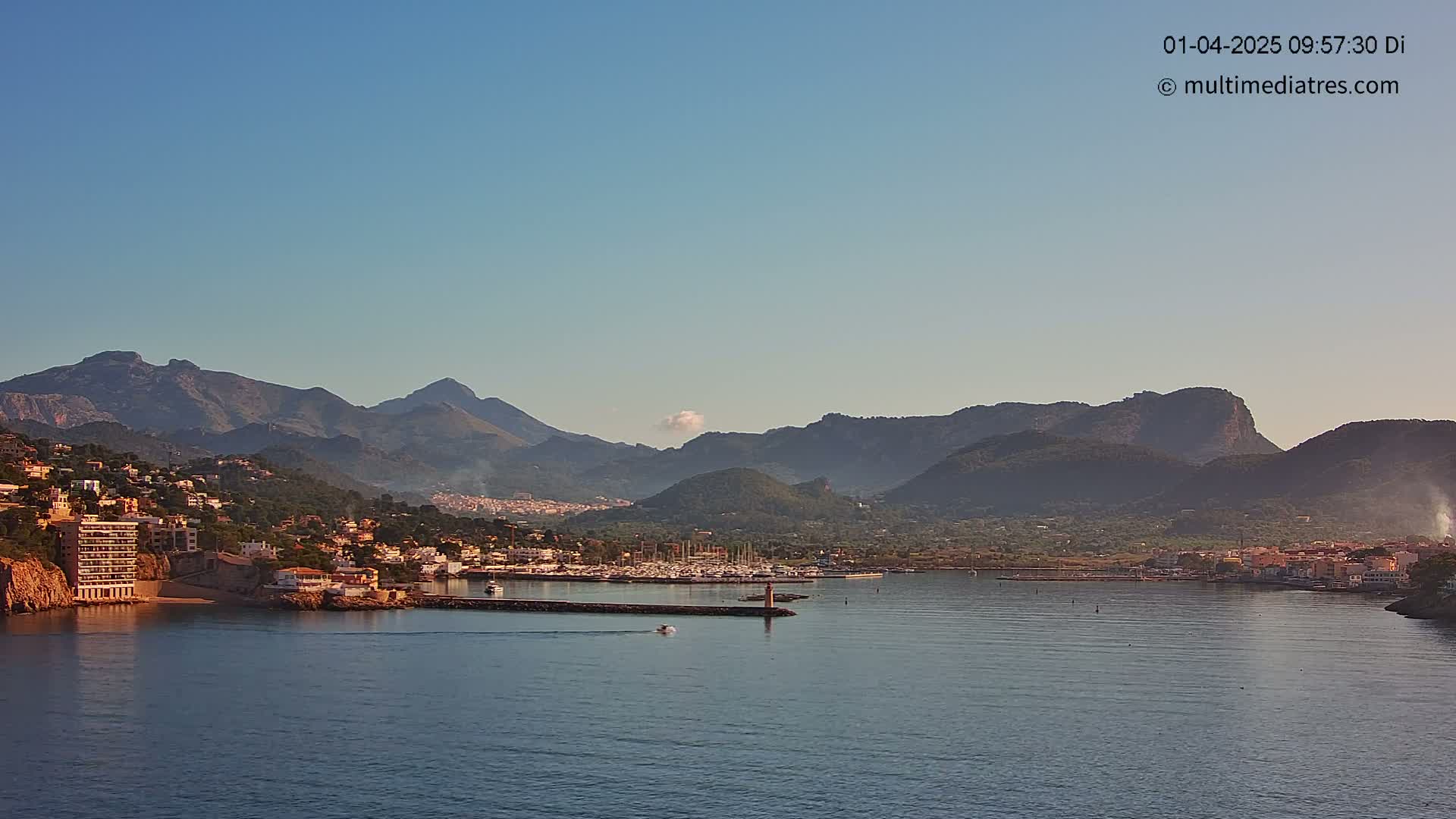 A coastal town nestled at the base of mountains under a clear blue sky is seen from across a calm body of water.