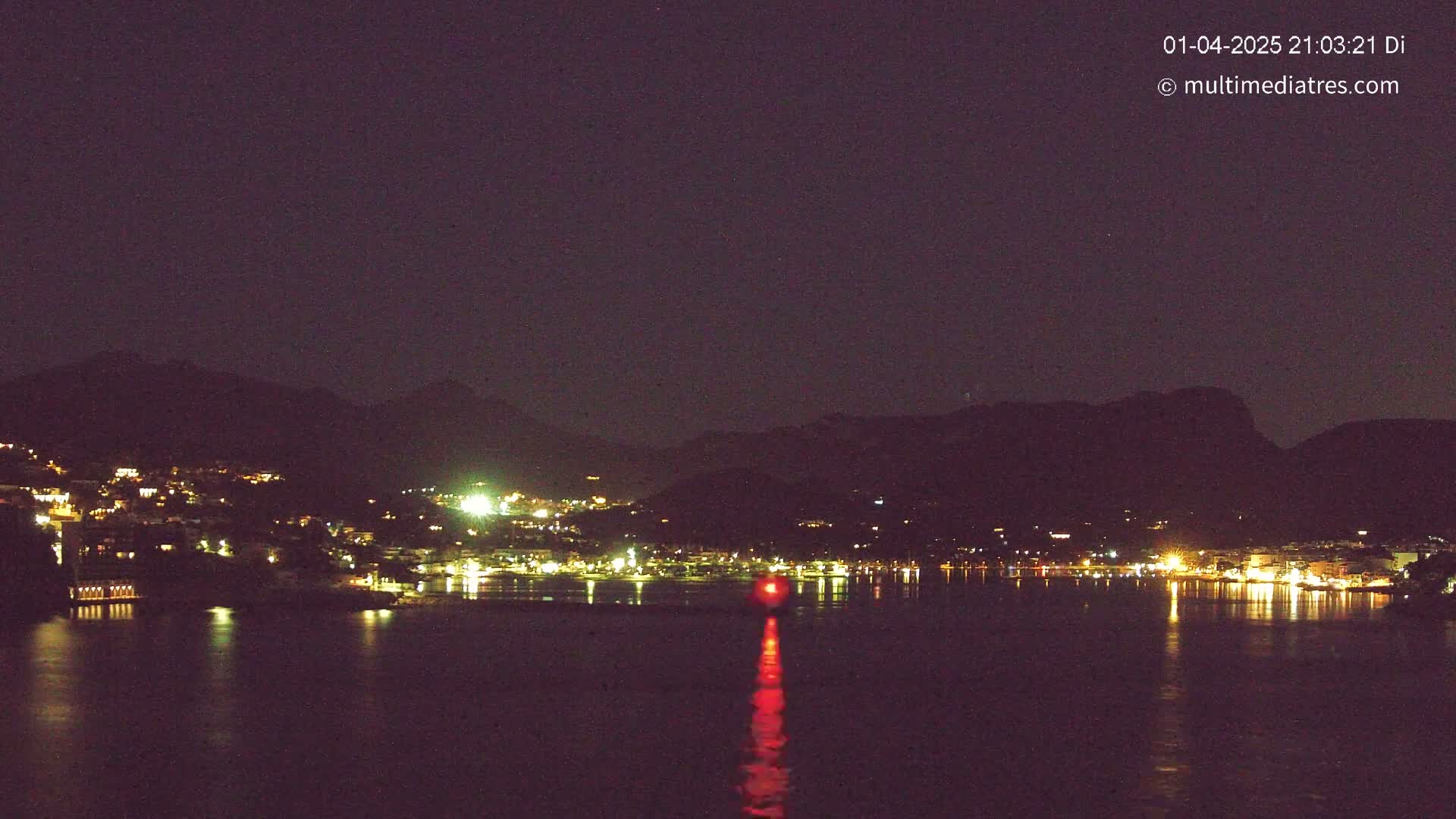 A nighttime view of a town's lights illuminating a dark body of water, nestled between silhouetted mountains under a clear, dark sky.