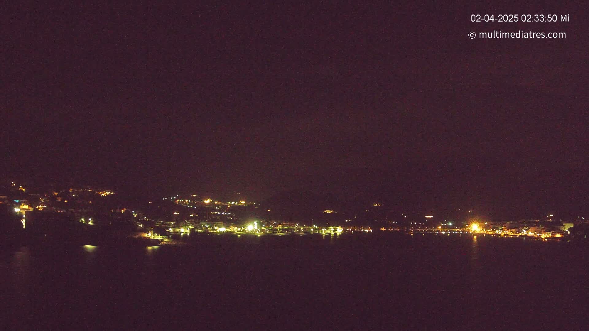 A nighttime view of a coastal town with scattered lights across a dark hillside, reflected in a still body of water under a clear, dark sky.