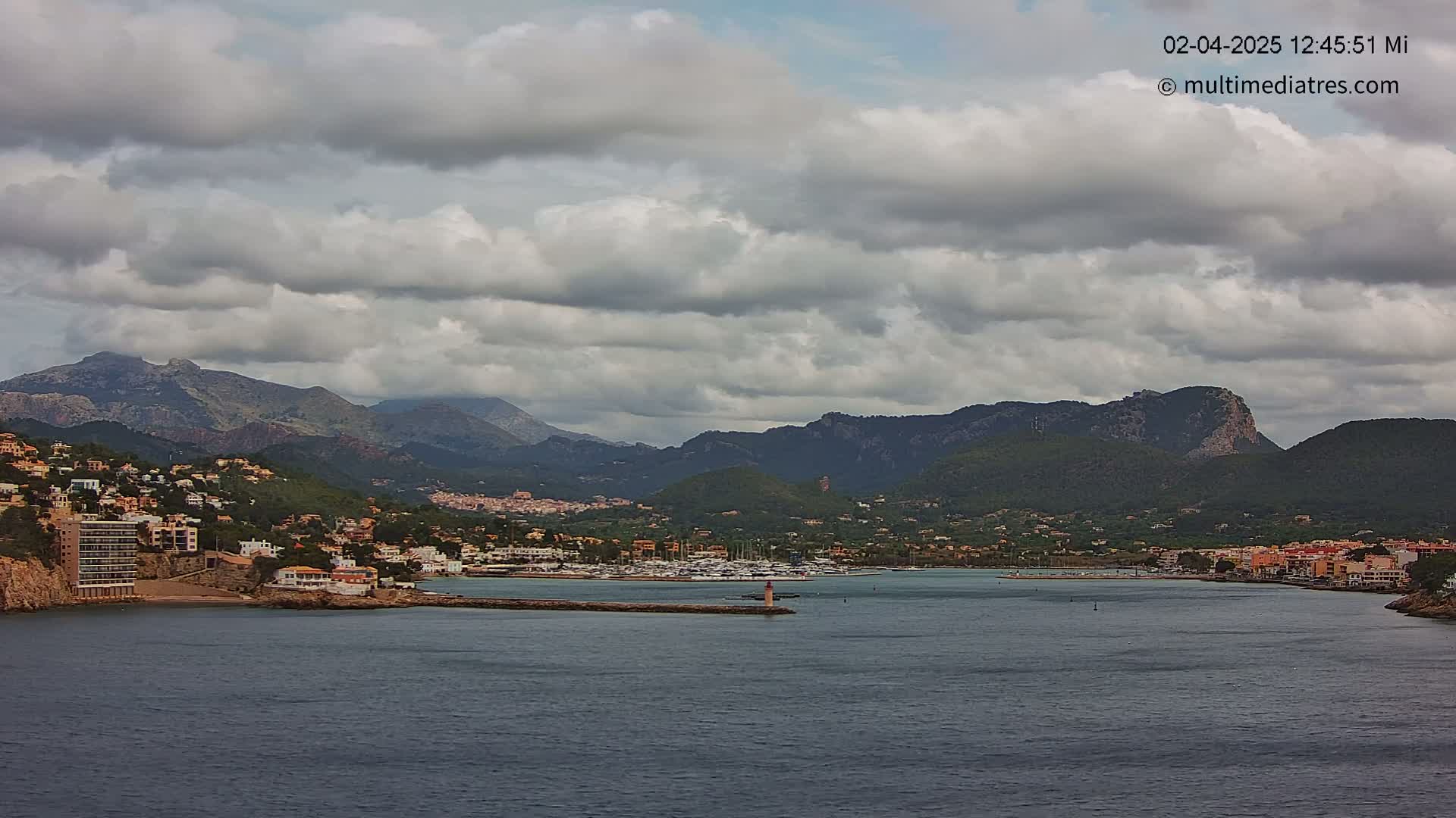 A coastal town nestled in a mountainous region under a cloudy sky sits on a bay with many boats.
