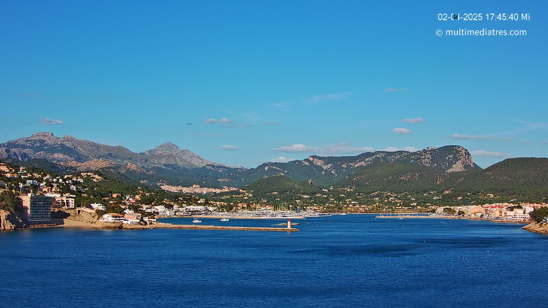 A coastal town nestled against a mountain range under a clear blue sky, with a calm blue sea in the foreground and numerous boats docked in a harbor.