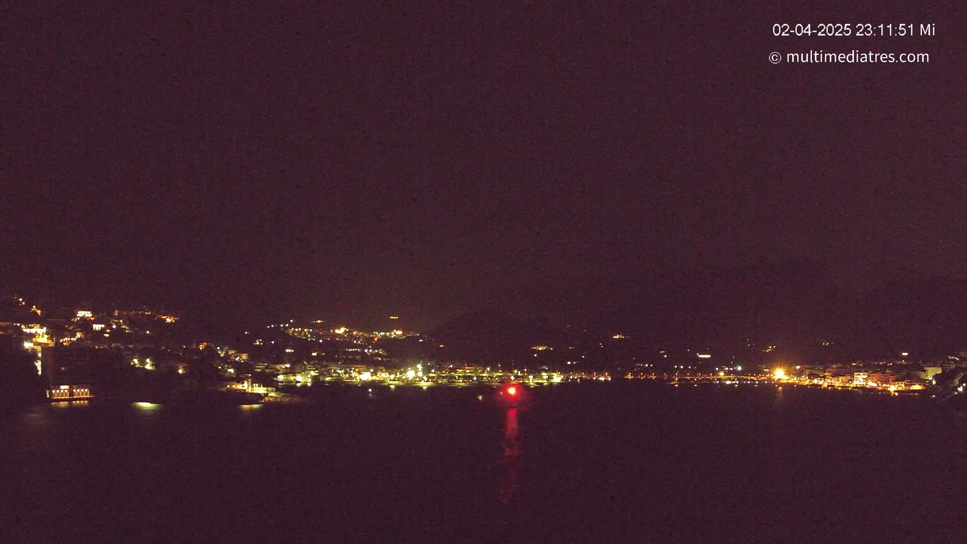 A nighttime, long shot of a coastal town illuminated by lights across a dark body of water under a clear, dark sky.