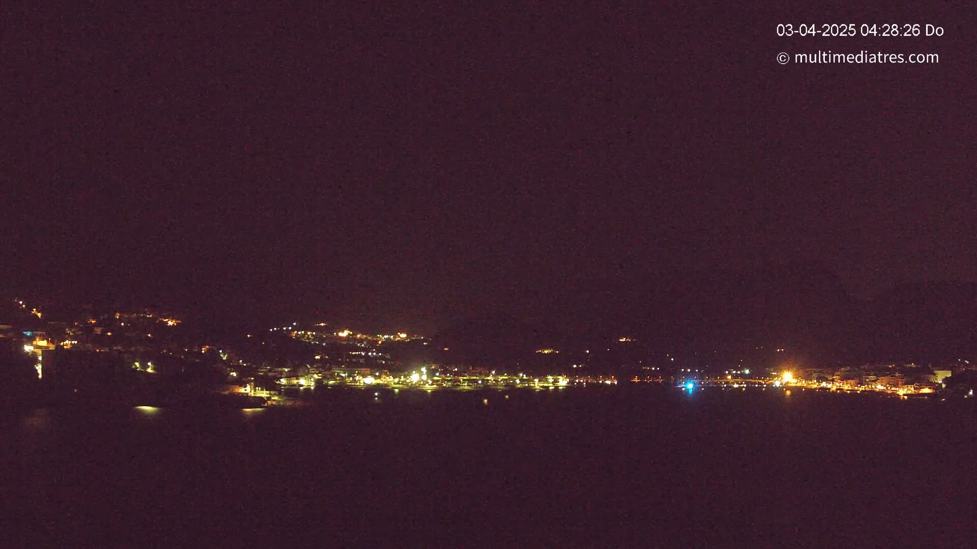 A nighttime view shows a town's lights scattered along a dark shoreline and hillside, with a calm, dark body of water in the foreground under a clear, dark sky.