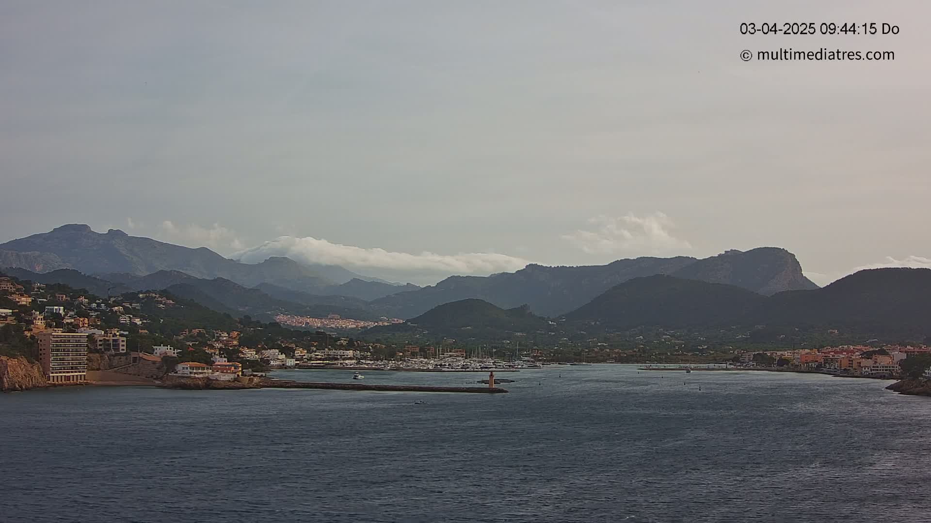 A coastal town nestled between dark mountains under a mostly cloudy sky overlooks a calm harbor filled with boats.