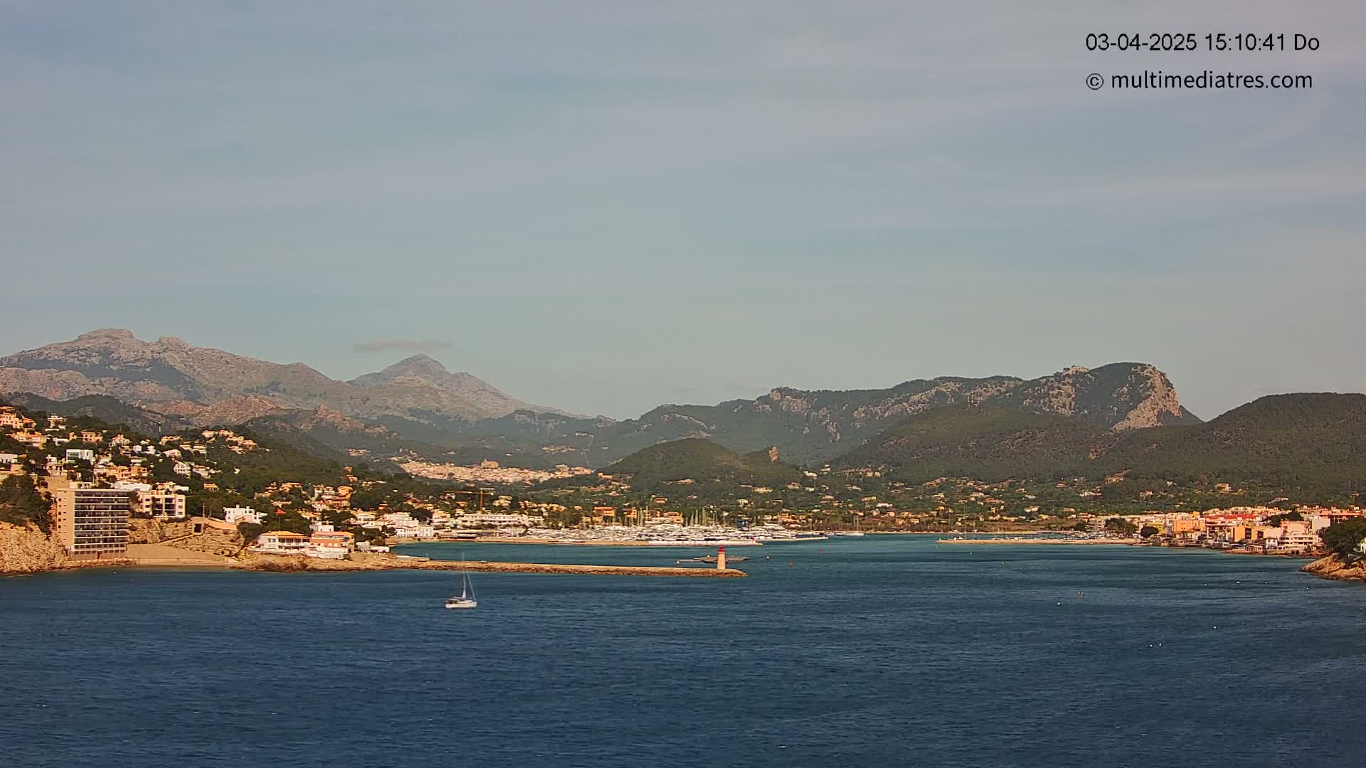 A coastal town nestled against a mountain range under a clear, light-blue sky, with a calm body of water in the foreground.