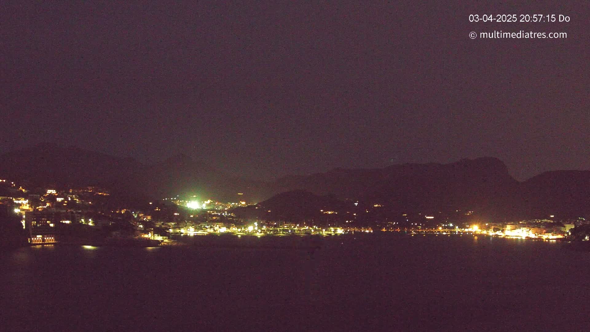 A nighttime view of a coastal town nestled between dark mountains, illuminated by scattered lights, under a clear, dark sky.