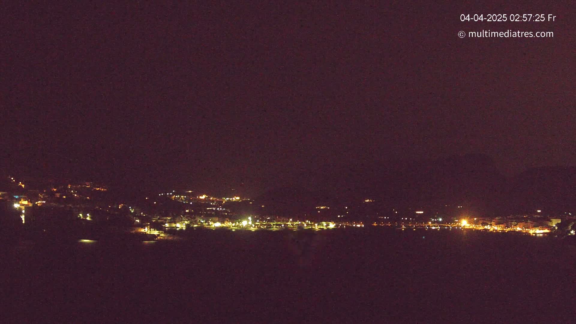 A coastal town at night, illuminated by scattered lights, is seen under a dark, clear sky.