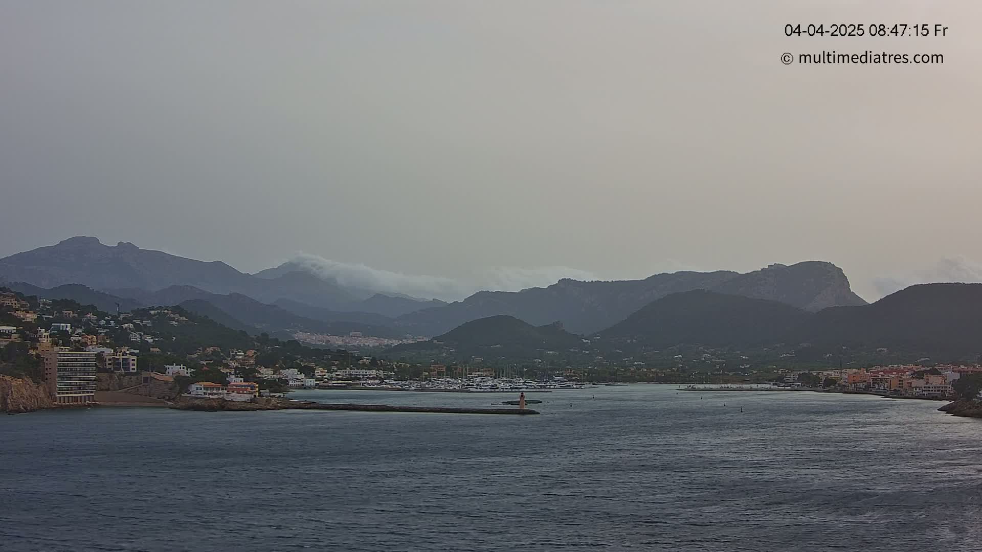 A hazy coastal town nestled at the base of mountains partially obscured by clouds, overlooking a calm body of water.