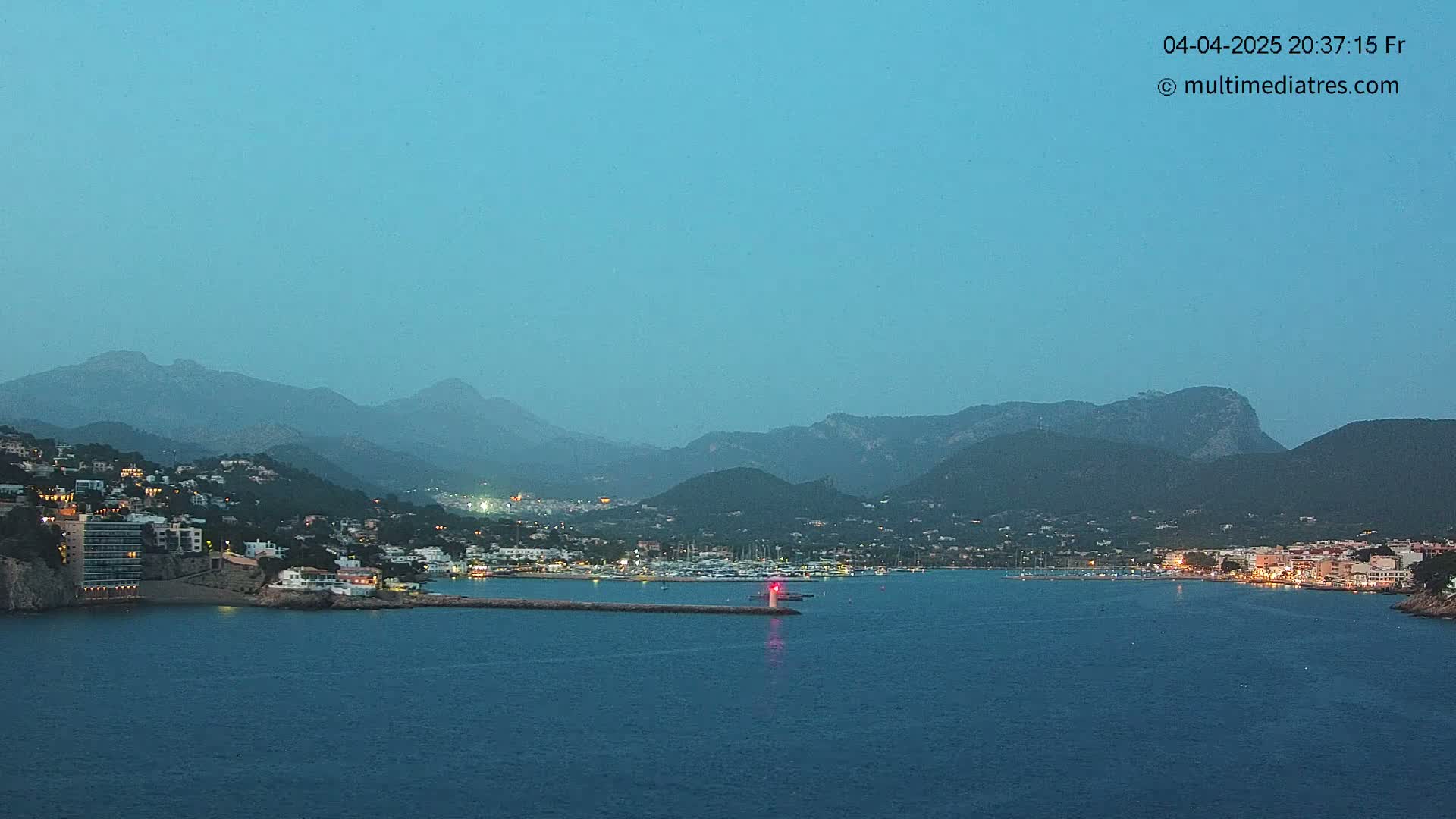 A coastal town at twilight, nestled between dark mountains and a calm, dark-blue sea, with a small harbor containing several boats.