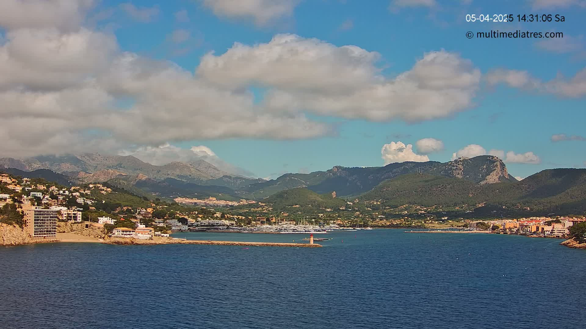 A partly cloudy day reveals a coastal town nestled between mountains and a calm blue sea, with a marina filled with boats.
