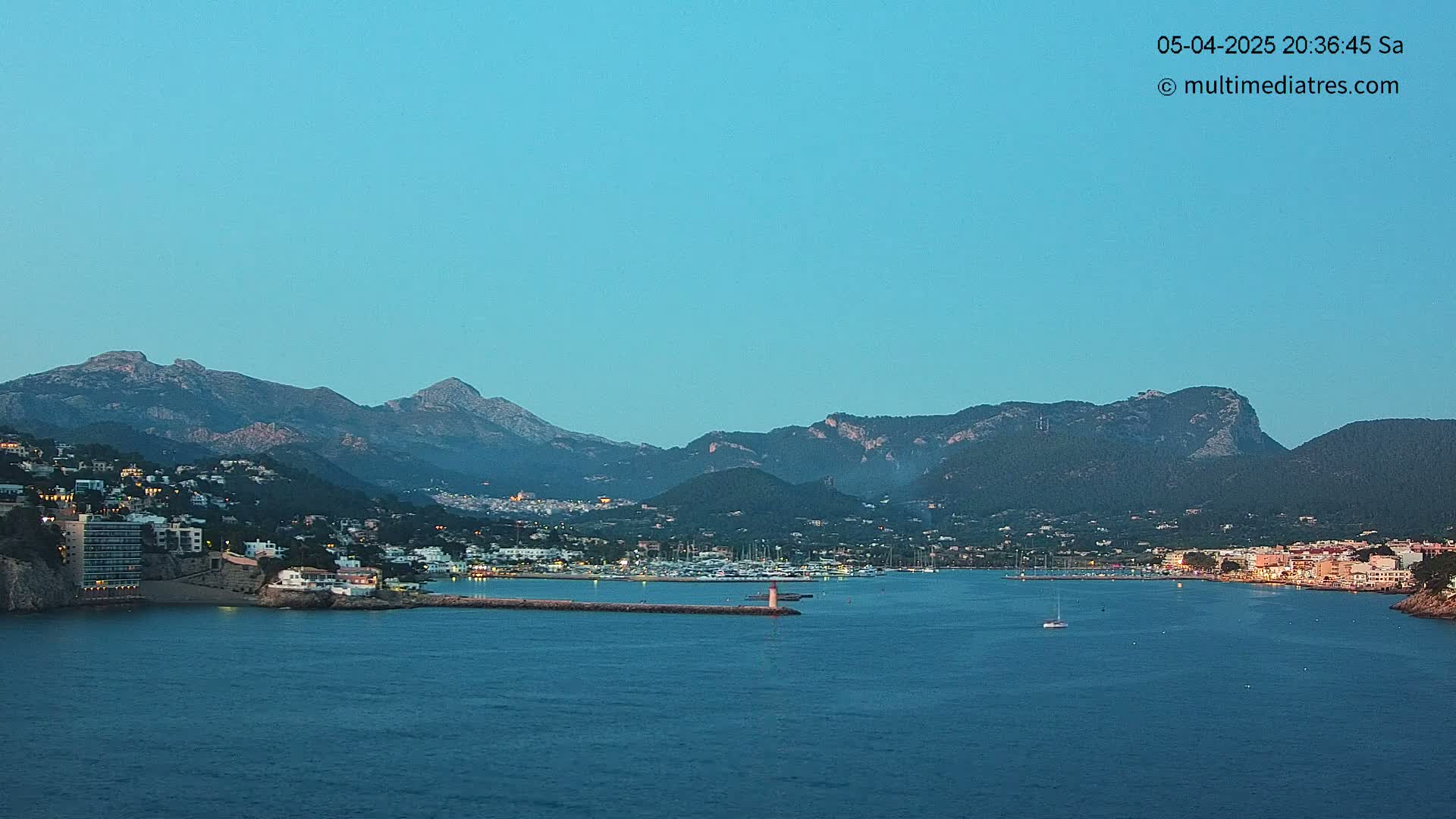 A coastal town nestled beneath mountains, with a calm harbor filled with boats, under a clear twilight sky.