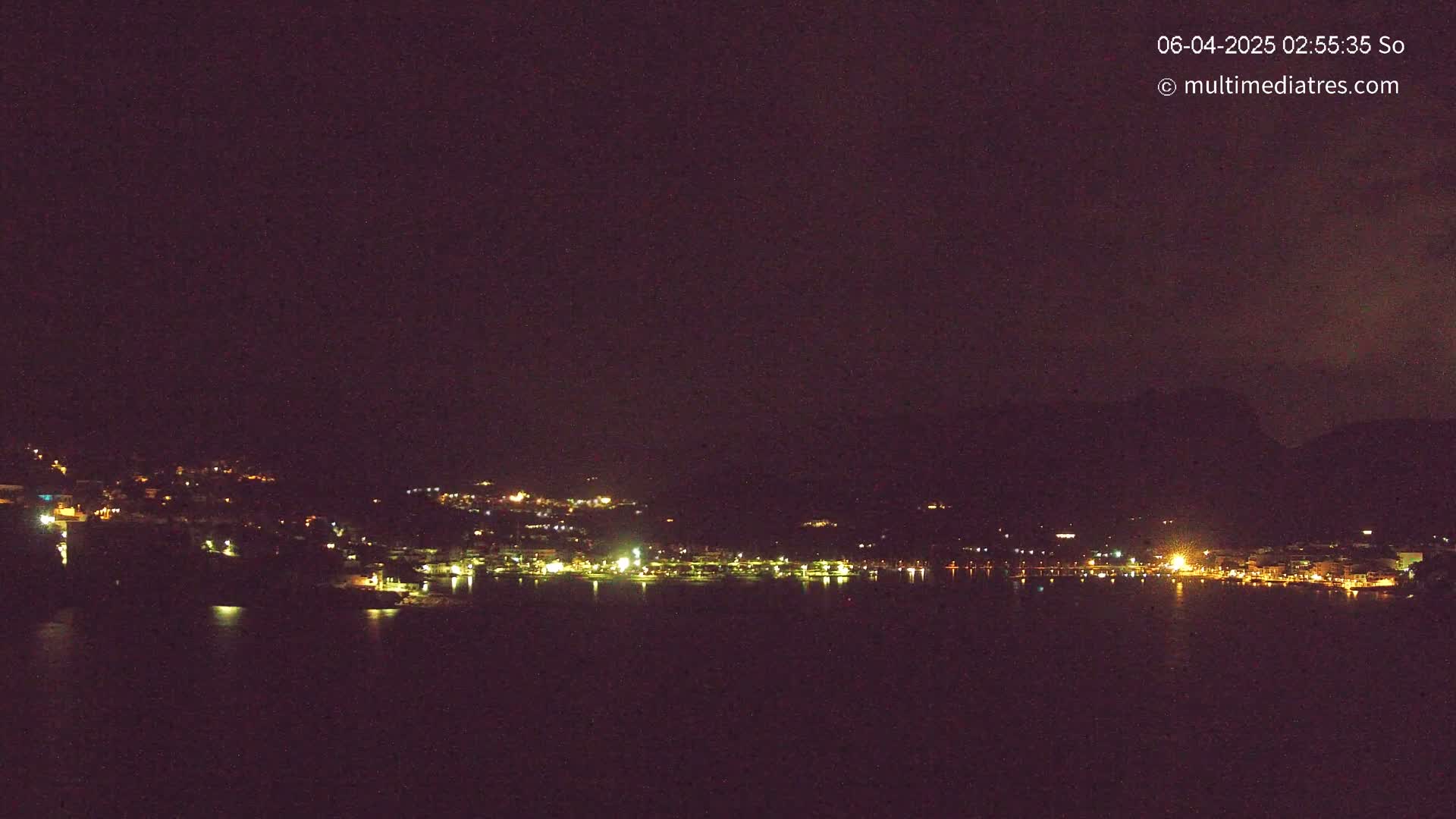A nighttime view of a town's lights across a dark body of water, nestled against a hill under a clear, dark sky.