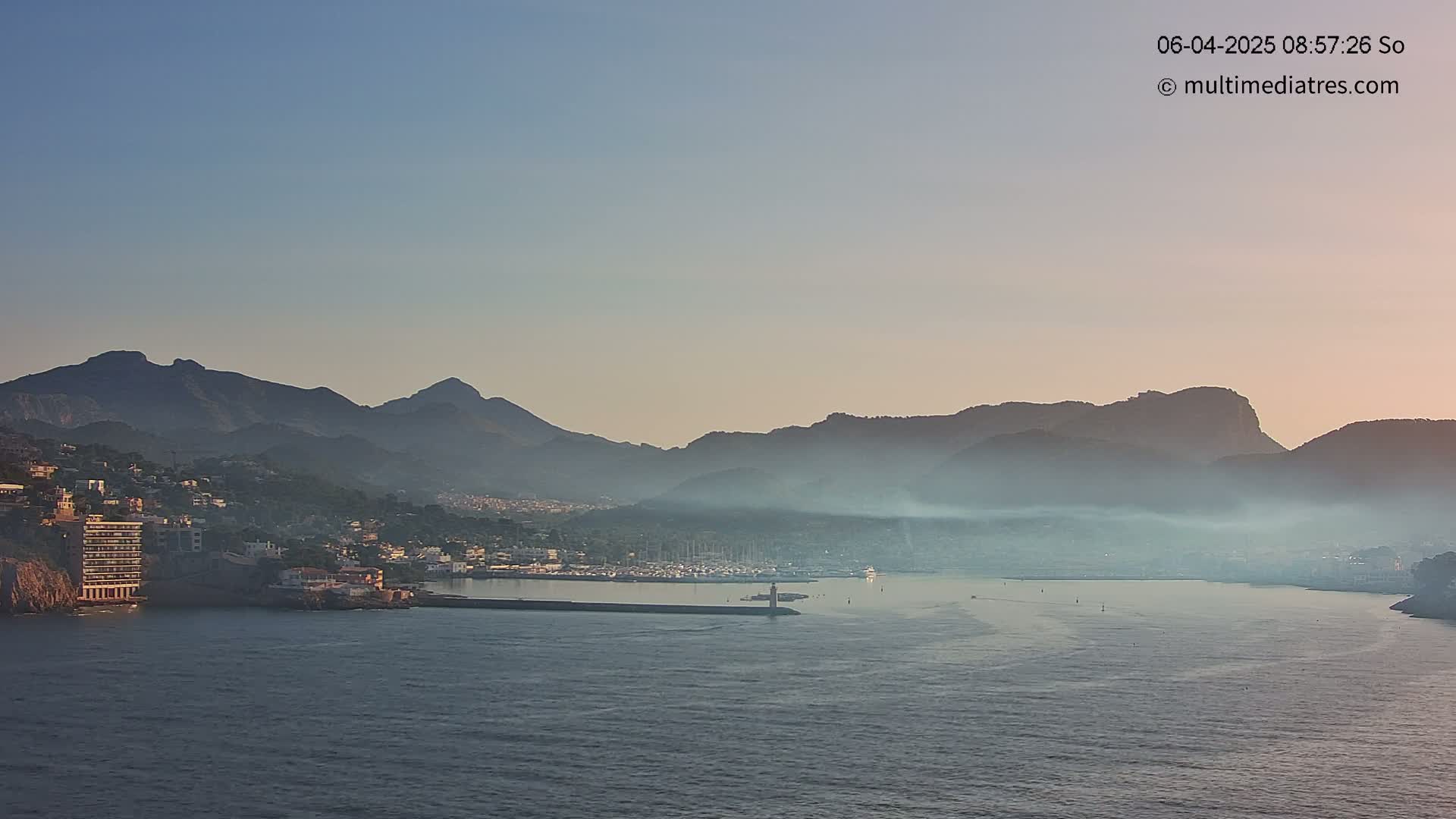 A hazy sunrise illuminates a coastal town nestled between a calm sea and a range of mountains.
