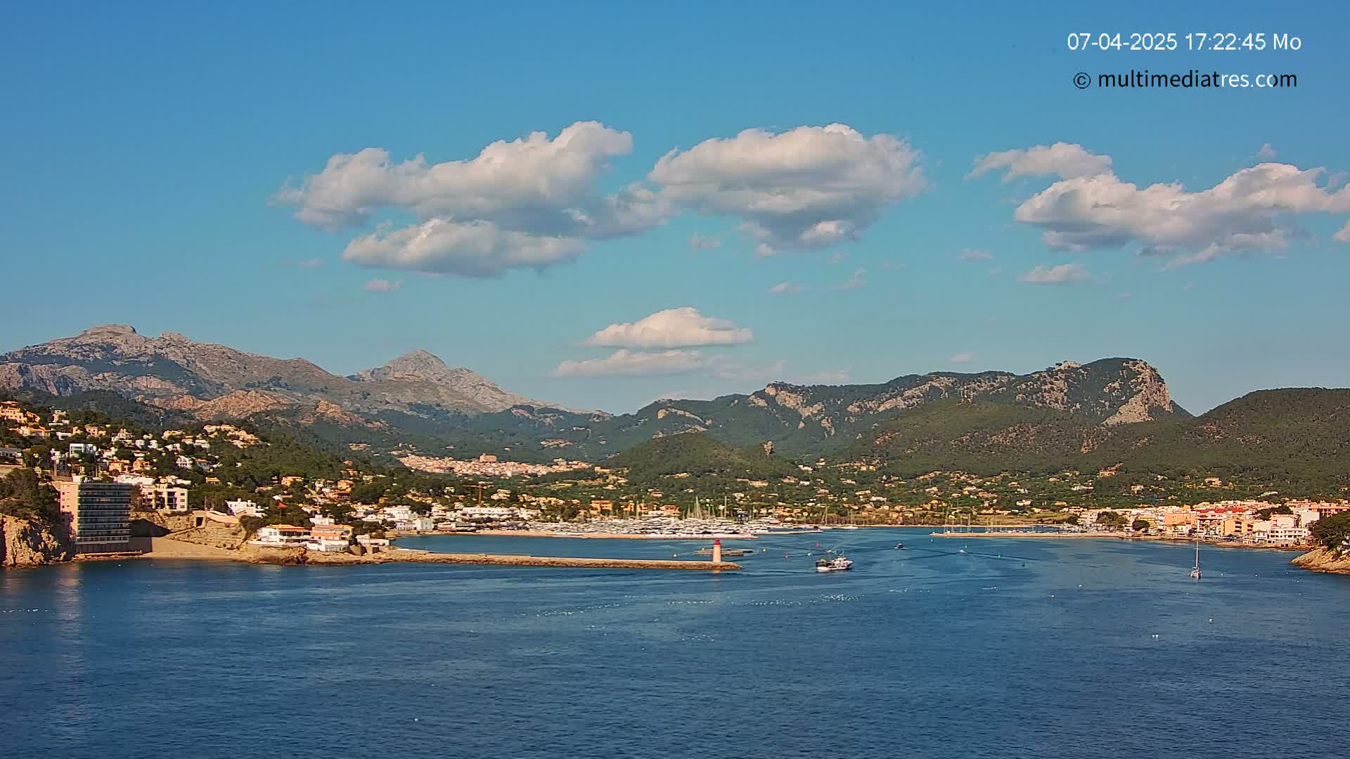 A coastal town nestled against a mountainous backdrop under a mostly sunny sky with scattered cumulus clouds.