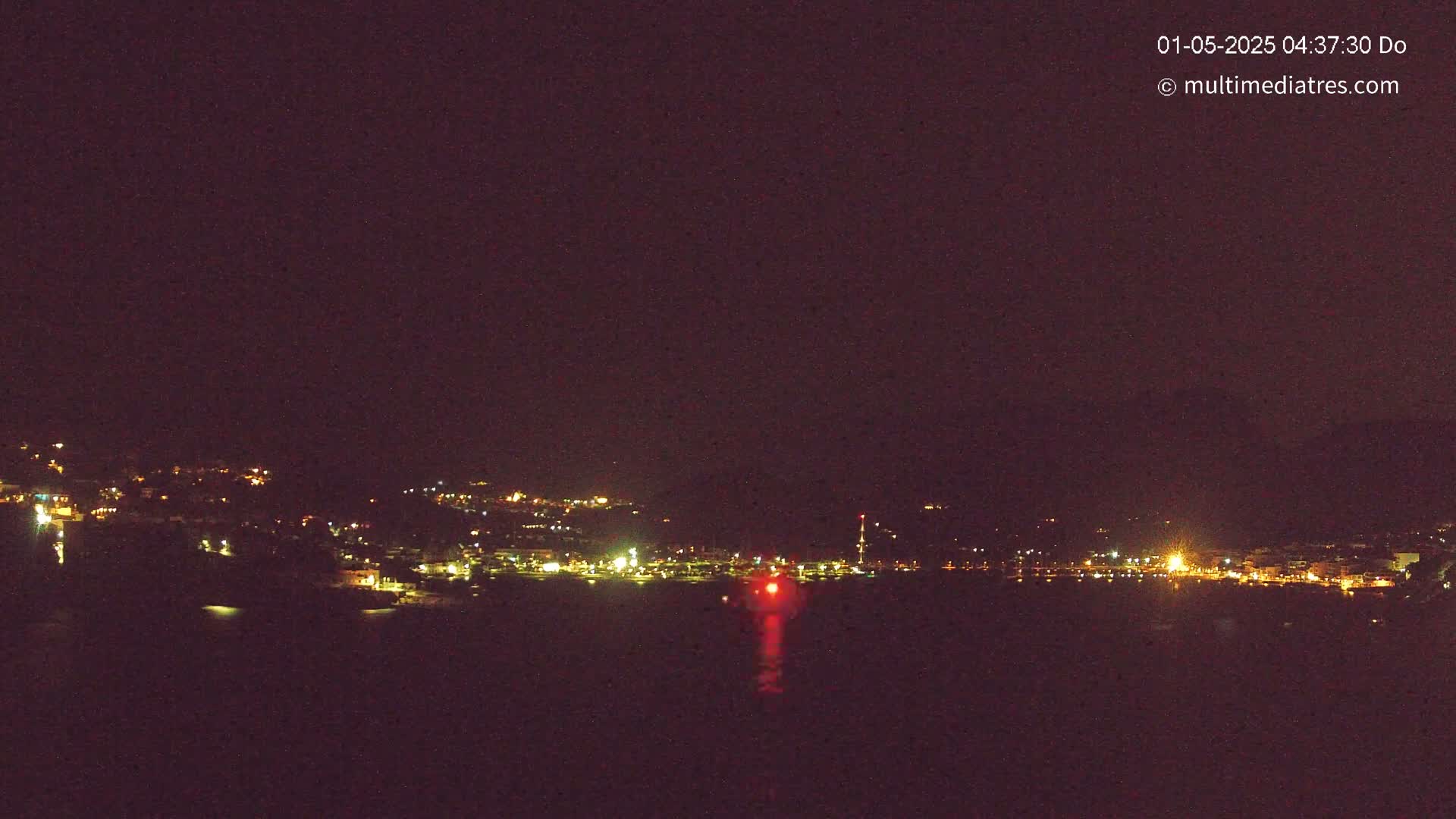 A nighttime long shot shows a town's lights spread across a hillside and reflecting in dark water.