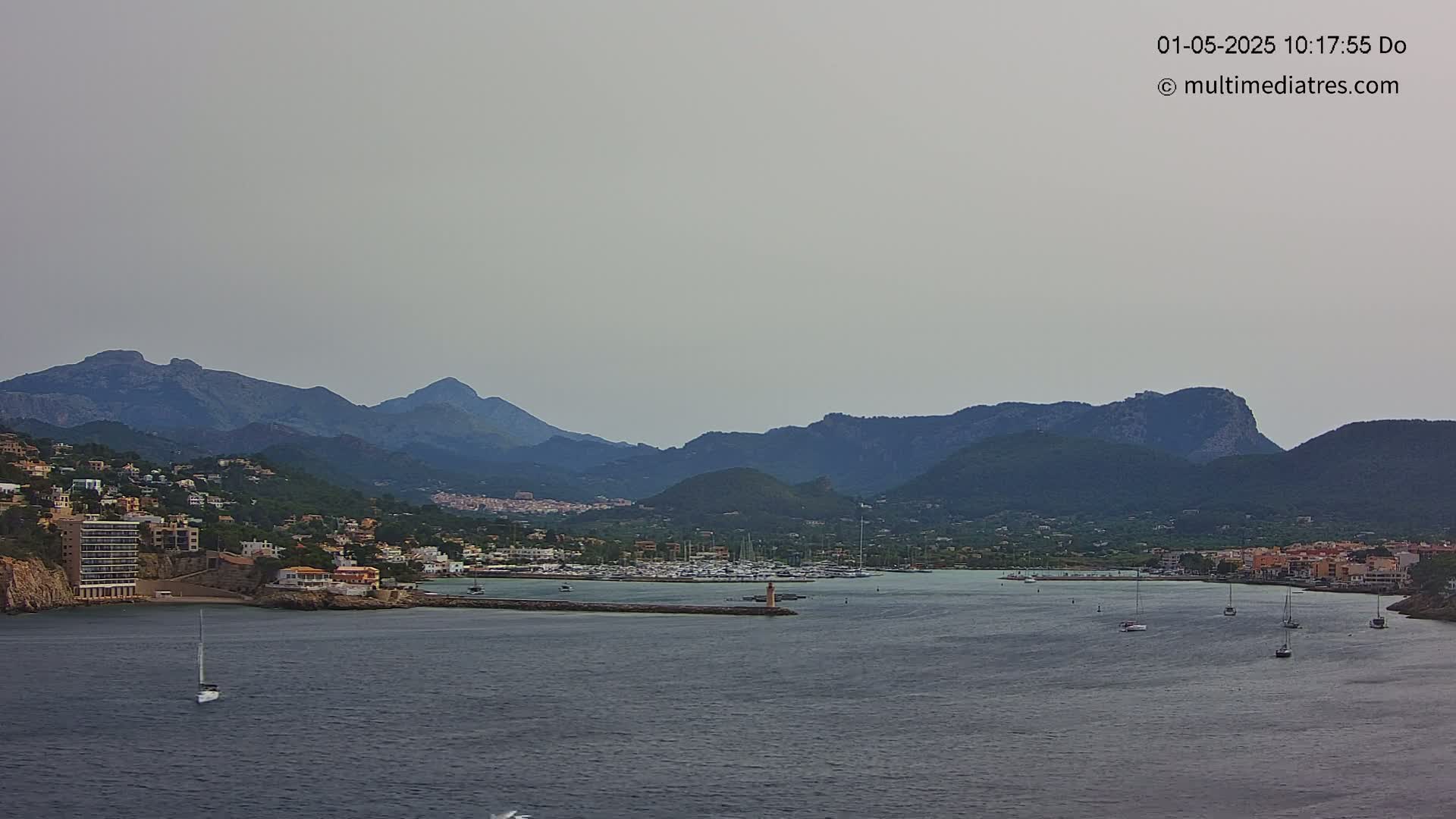 A hazy view shows a calm bay with several sailboats, a small marina, and a town nestled against mountains under a muted sky.