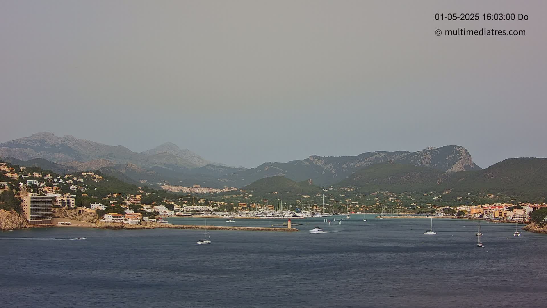A hazy coastal town nestled against a mountain range, with a calm bay containing several boats.