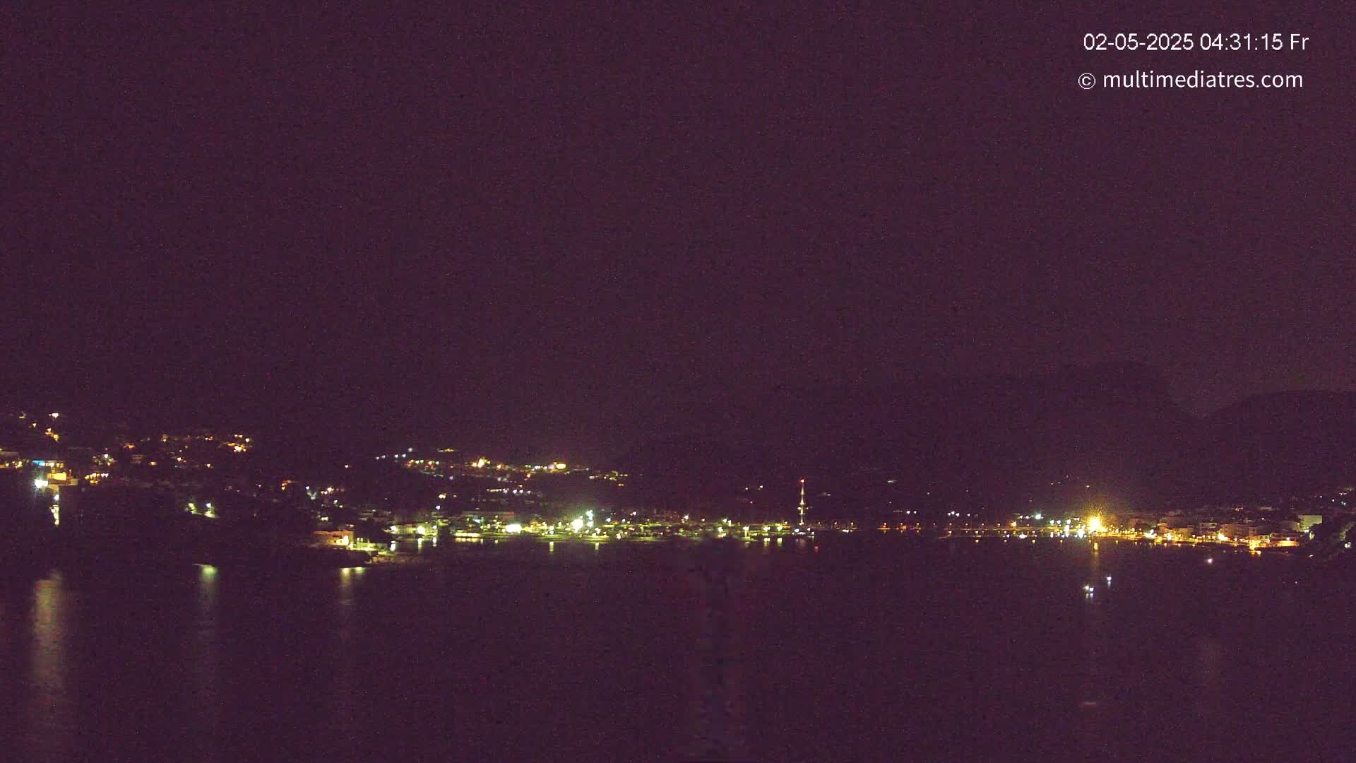 A nighttime long shot shows a town's lights spread across a hillside beside a dark body of water under a clear, dark sky.