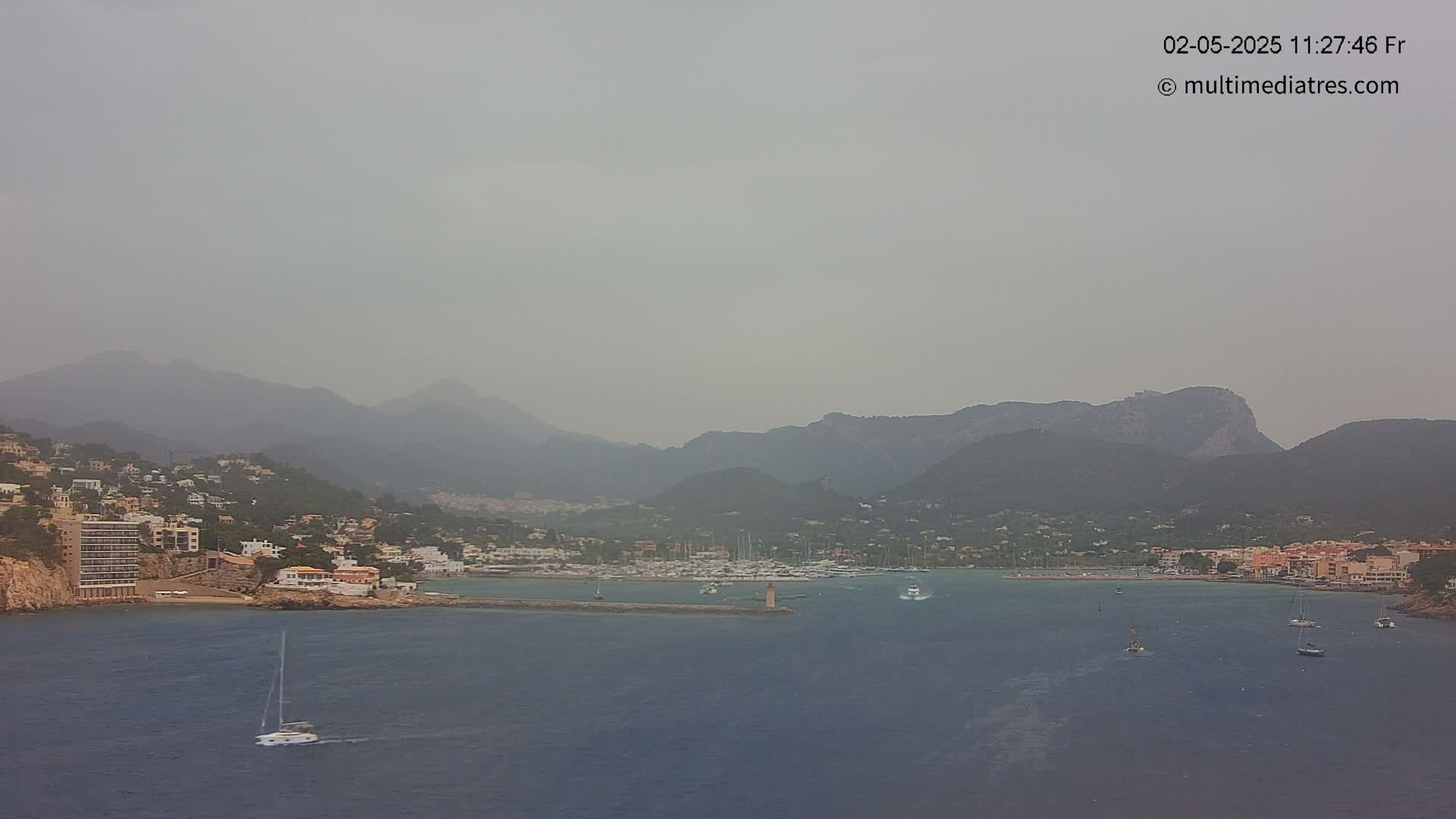 A hazy view of a coastal town nestled between mountains and a calm body of water with several sailboats.