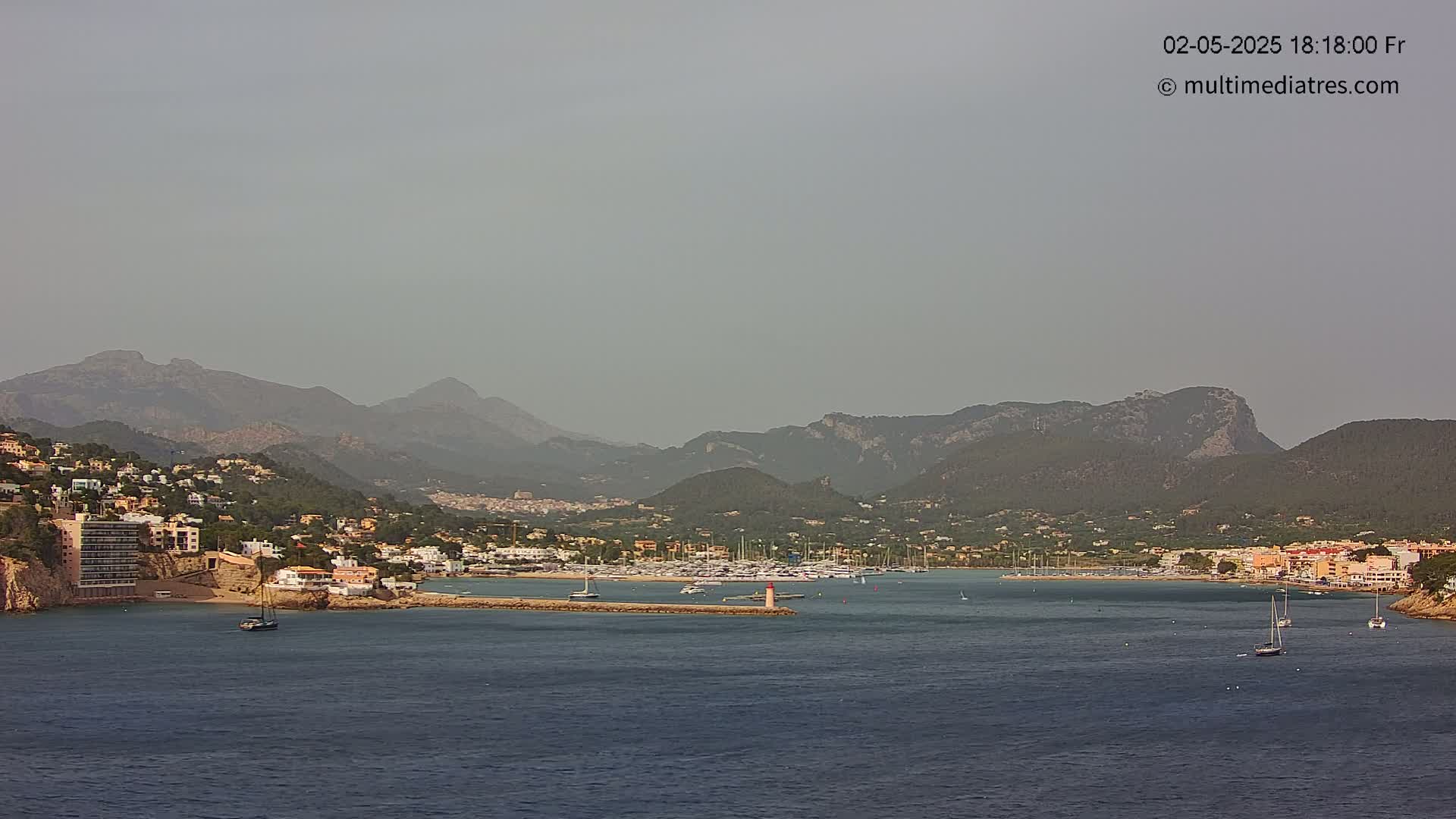A hazy coastal town with a marina nestled between dark green mountains and a calm body of water.