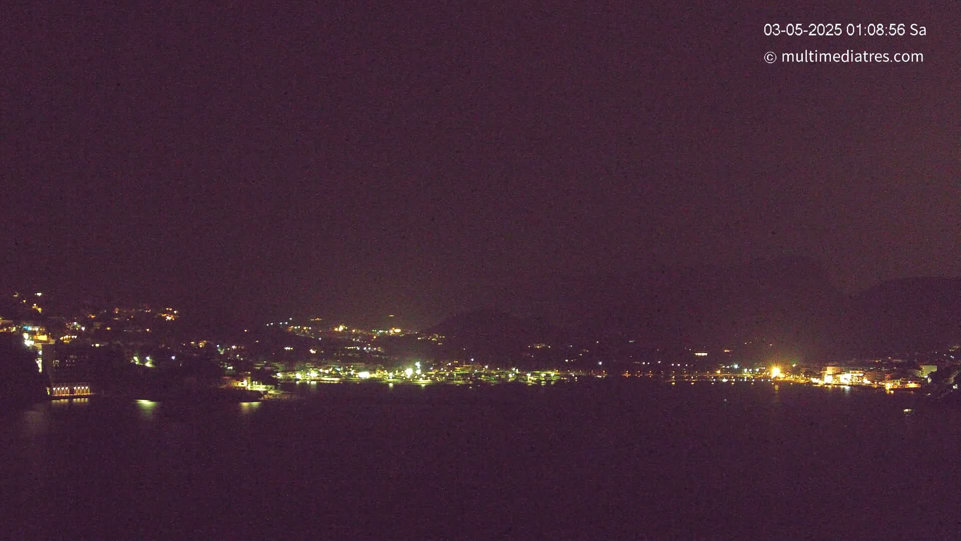 A nighttime view shows a dark, calm body of water reflecting the scattered lights of a town nestled on a hillside under a clear, dark sky.
