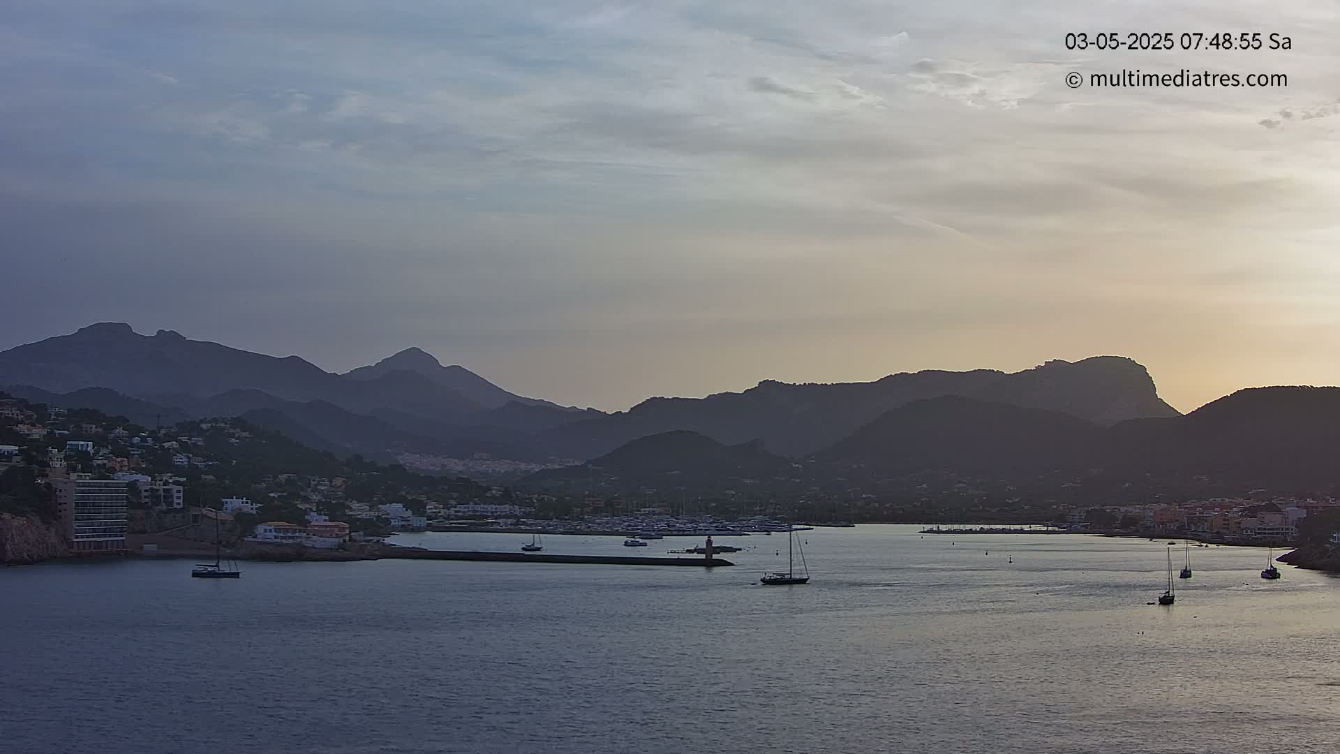A calm bay with several sailboats is nestled between a coastal town and a range of dark mountains under a partly cloudy, dusky sky.