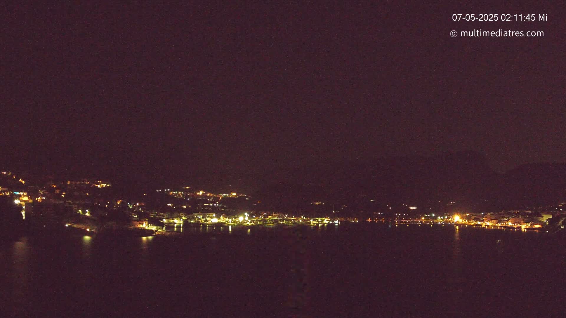 A nighttime view of a coastal town with many lights, seen from across a dark body of water under a clear, dark sky.