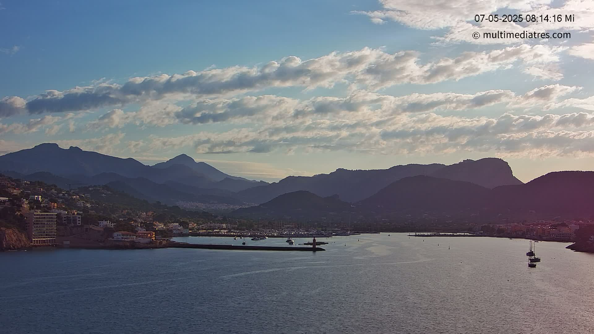 A coastal town nestled at the base of a mountain range under a partly cloudy sky is seen from across a calm body of water.