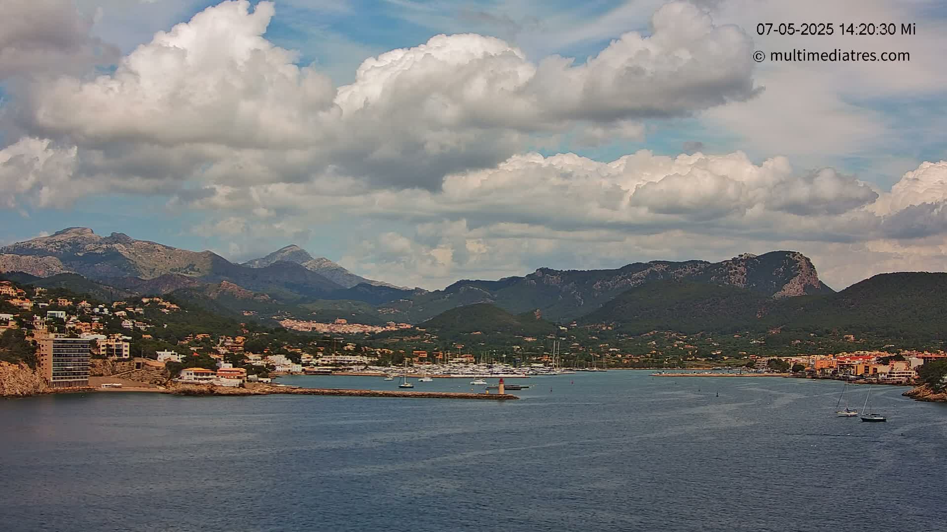 A partly cloudy day reveals a coastal town nestled between mountains and a calm harbor filled with boats.