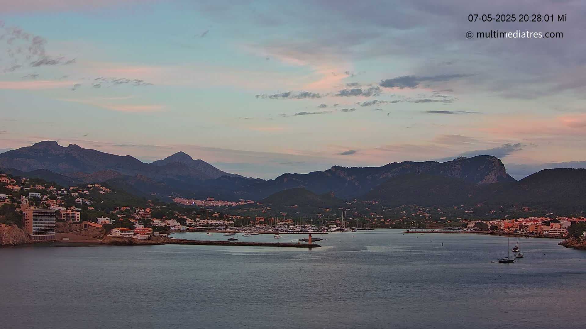 A calm harbor with a small marina is nestled between a town and a range of dark mountains under a pale pink and purple twilight sky.