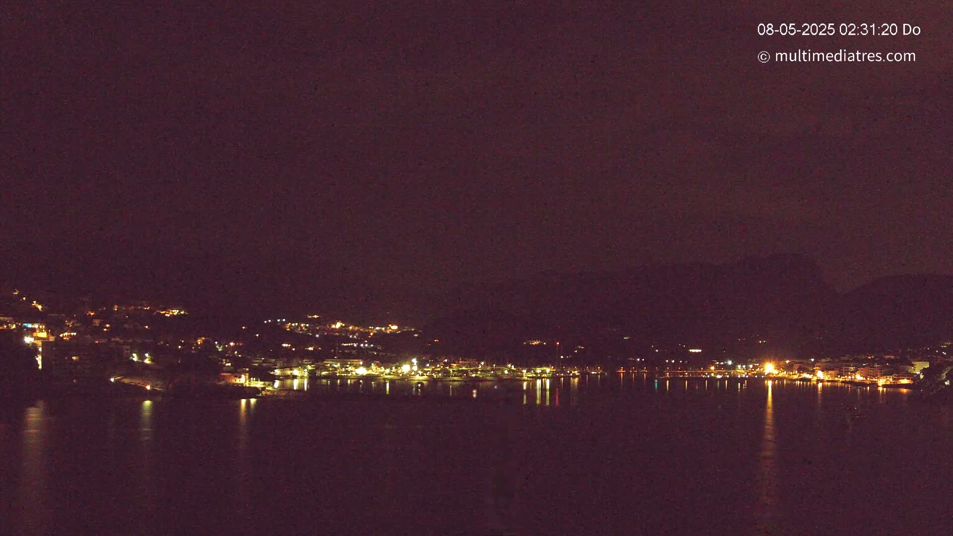 A nighttime view shows a dark, calm body of water reflecting the lights of a town nestled against a hillside under a clear, dark sky.